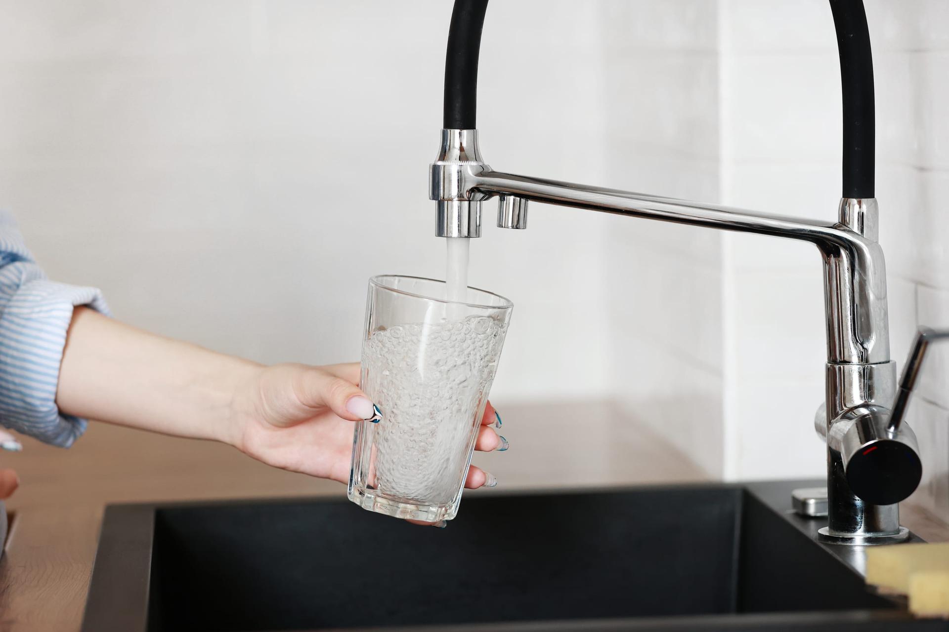 A hand holding a clear glass under a running chrome kitchen tap above a dark sink