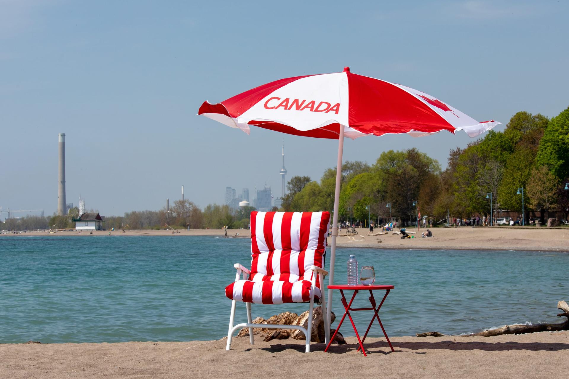 A striped deck chair under a red-and-white Canada parasol on a sandy Toronto beach, with Lake Ontario and the distant CN Tower behind.