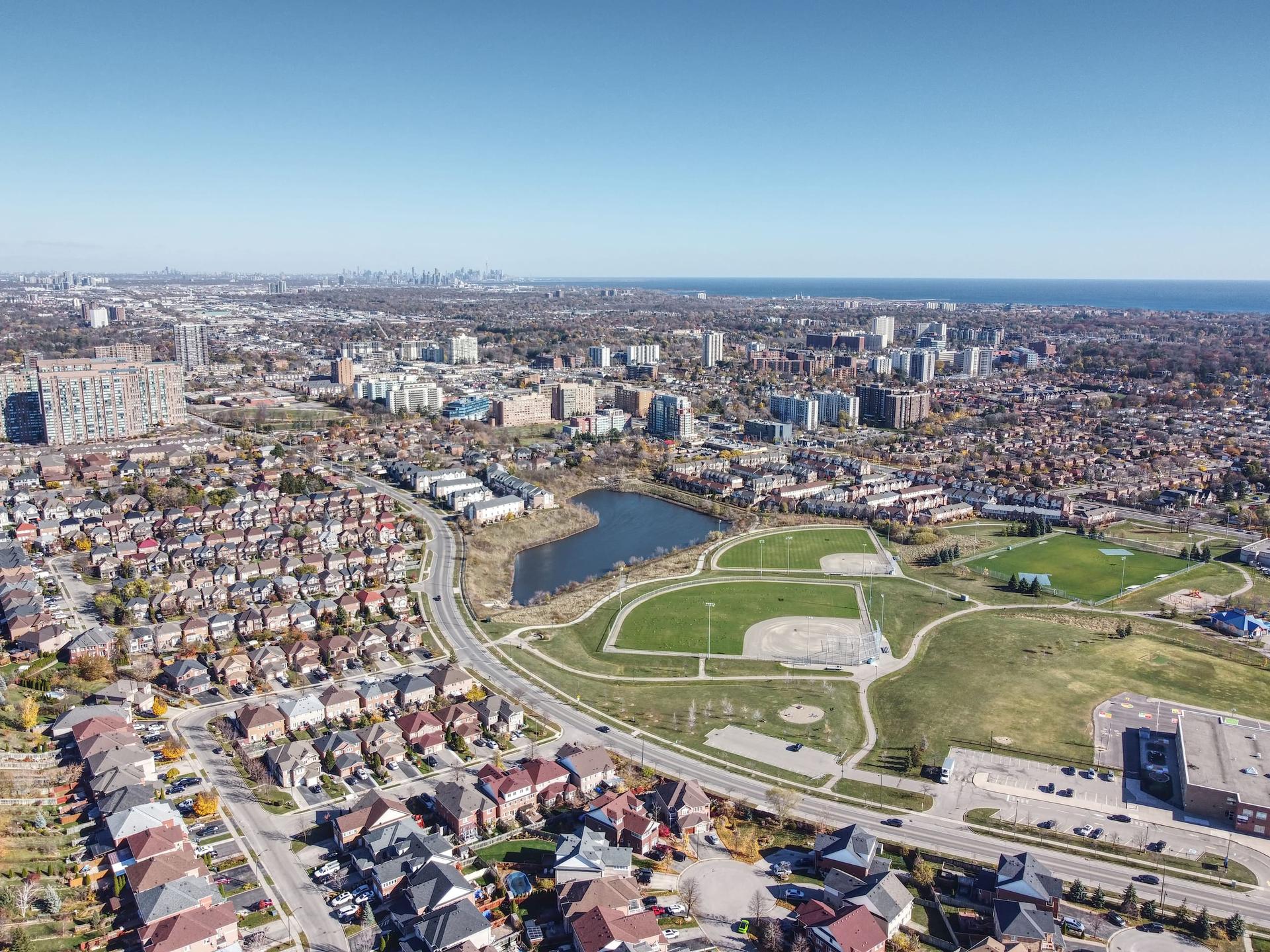 Aerial view over a Mississauga suburb: rows of detached houses and townhouses, a park with a pond and two ball diamonds, mid-rise and high-rise apartment towers, and Lake Ontario with the Toronto skyline on the horizon