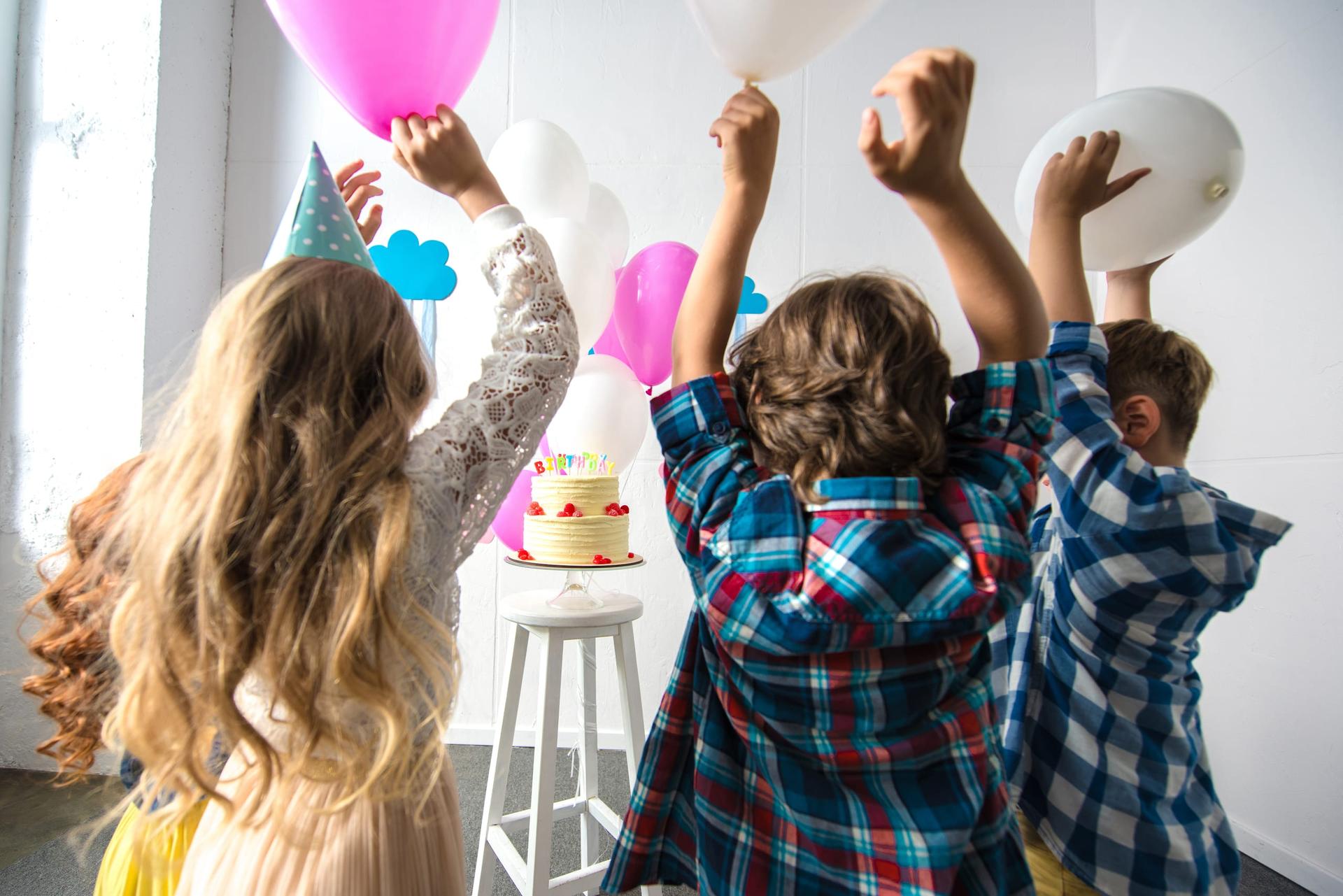 Three children holding balloons up beside a birthday cake on a stool