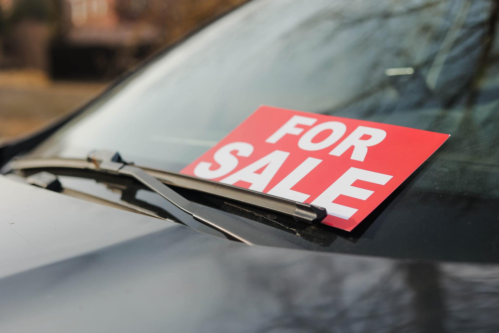 A red For Sale sign tucked under the windshield wiper of a parked car