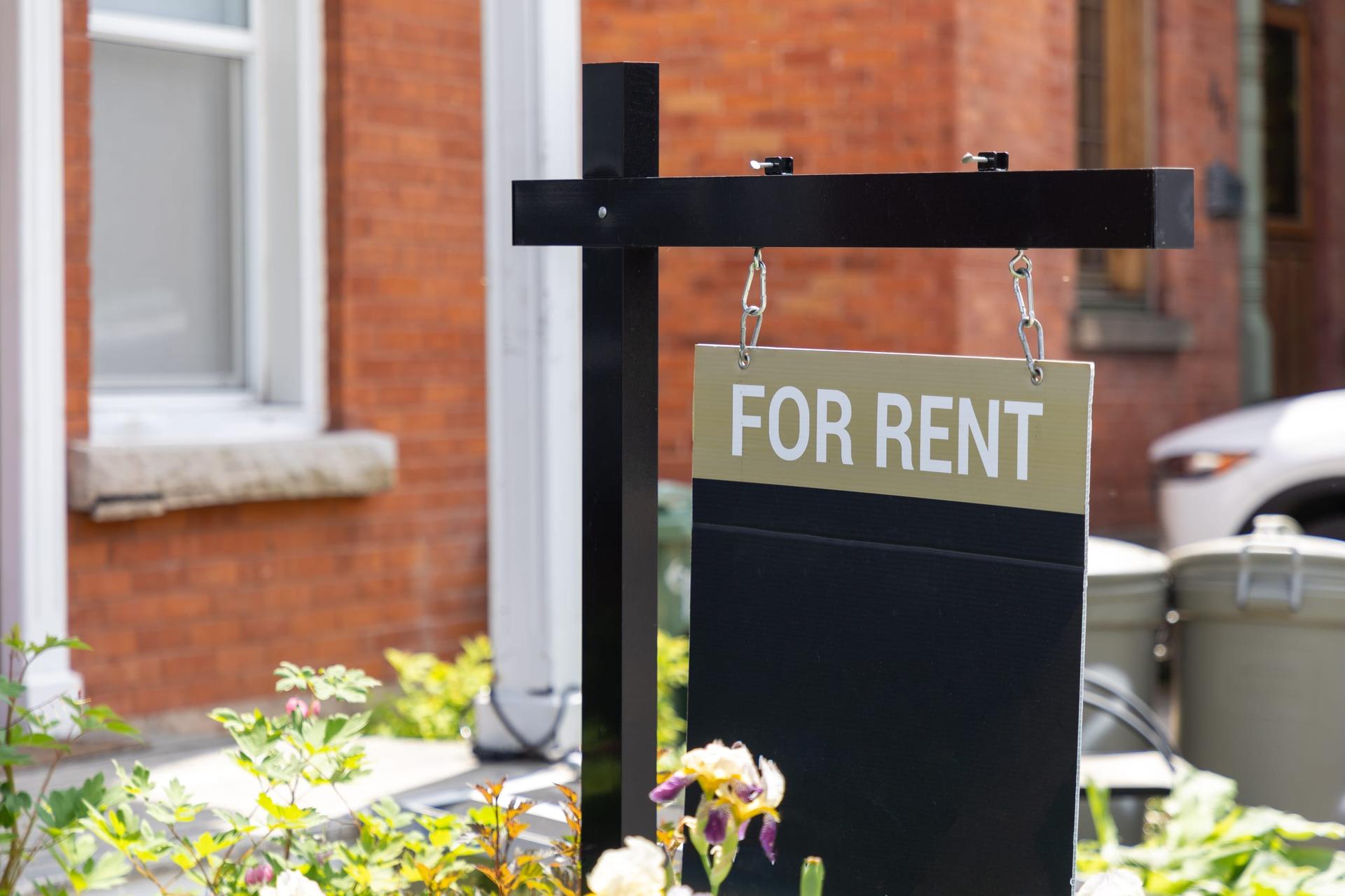A black and gold For Rent sign hanging from a post in front of a red-brick house with white window trim, with two wheeled waste bins beside the driveway