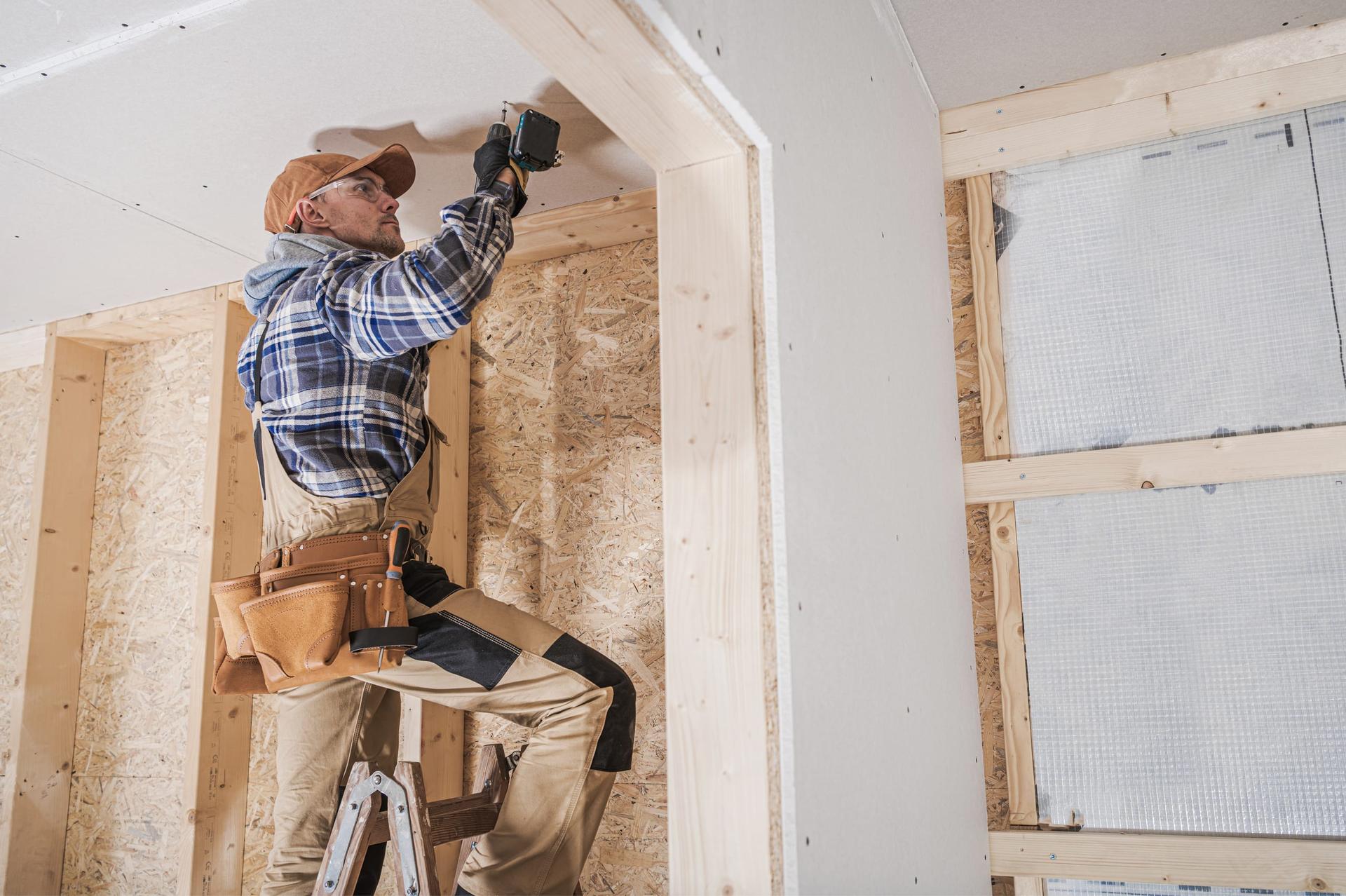 A contractor in a cap, safety glasses, plaid shirt and tool belt stands on a wooden stepladder and drives screws into a drywall ceiling beside a framed doorway, in a room with OSB-sheathed walls and exposed wood studs.