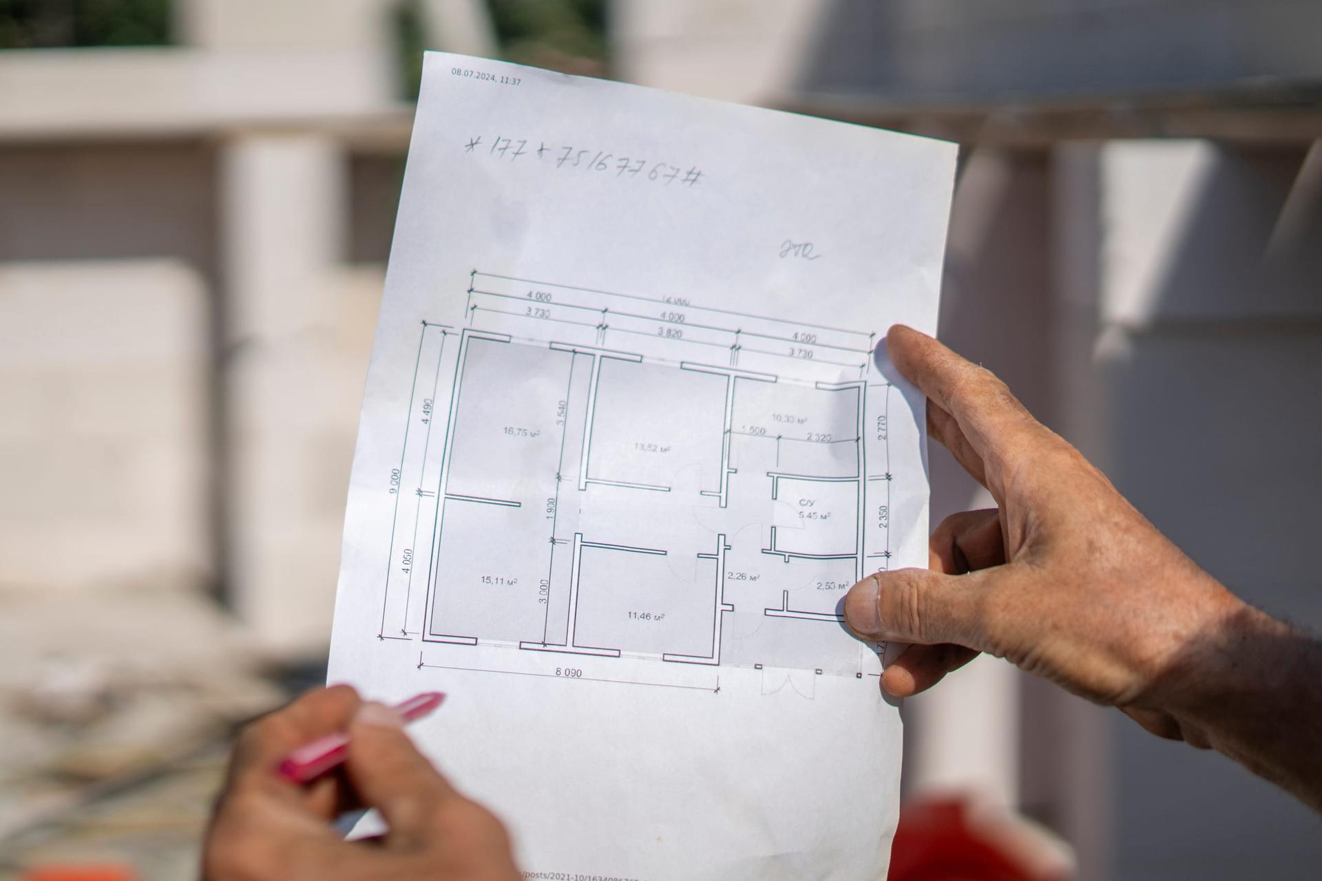 A construction worker holding an unrolled set of building plans in front of a house under construction.