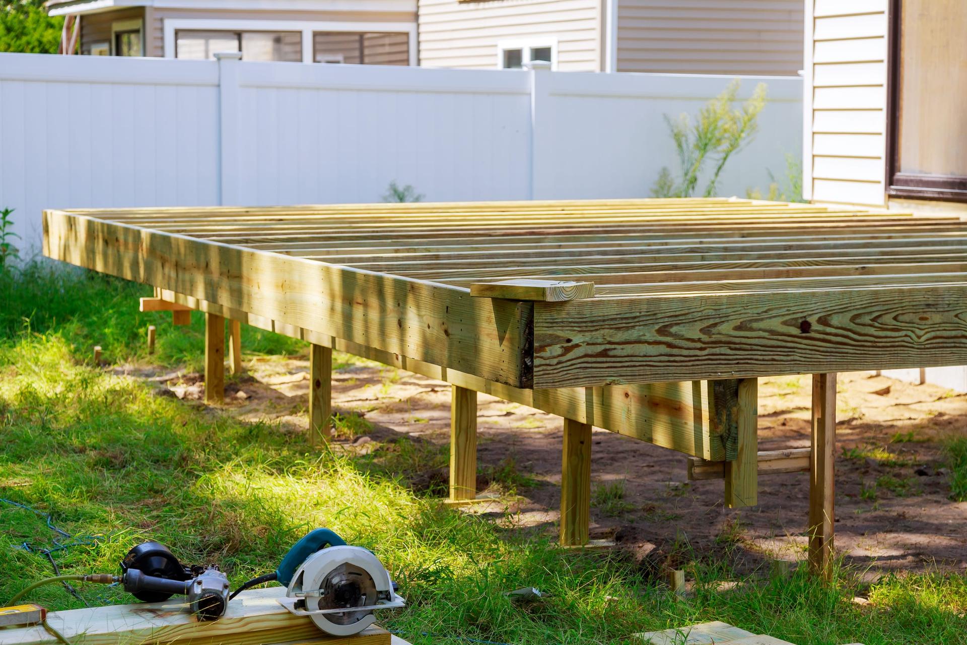 A raised deck frame of pressure-treated joists on posts in a fenced backyard, with a circular saw and ear defenders on a board in the foreground.