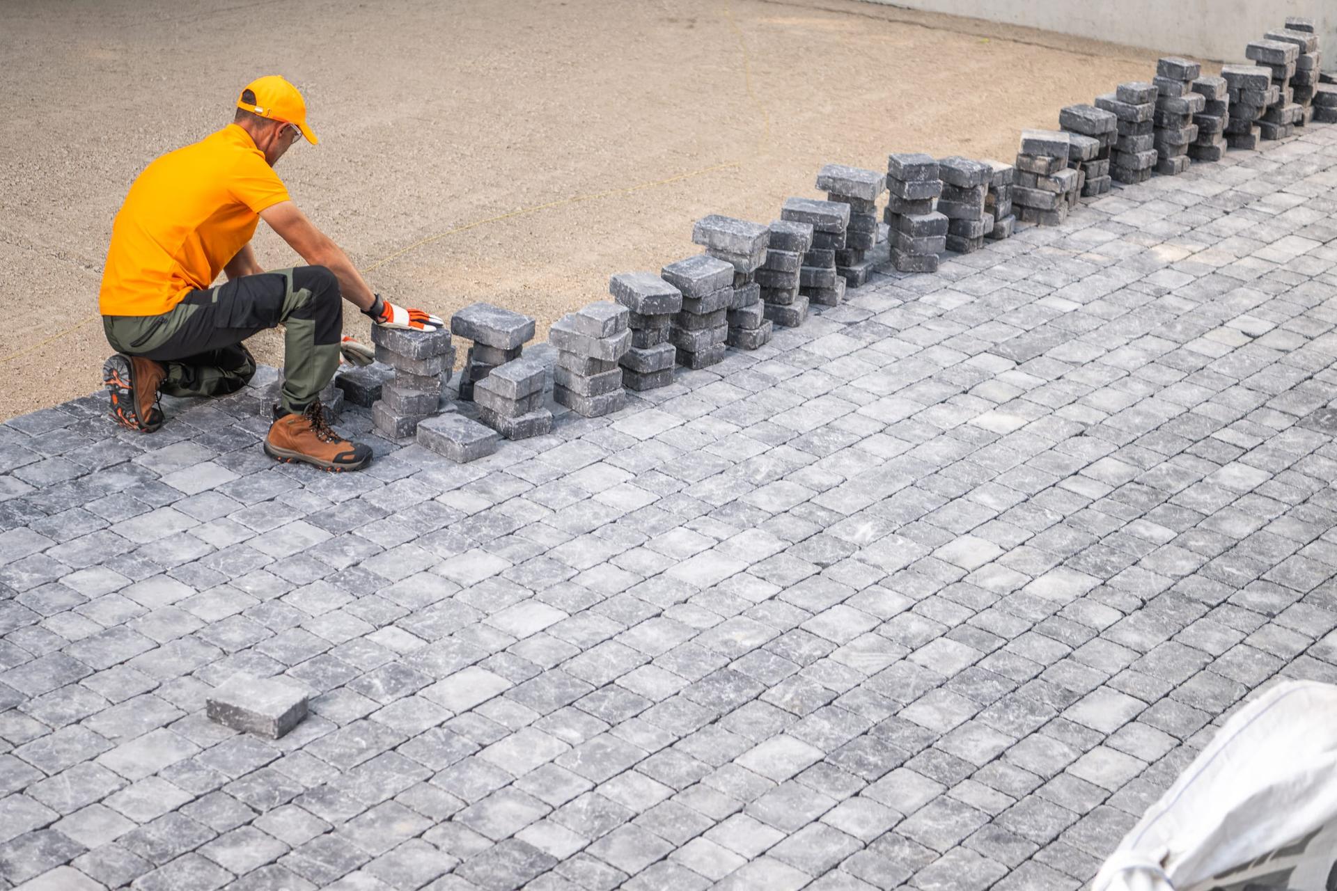 A worker in an orange shirt and hard hat kneeling to set grey interlocking pavers on a driveway, with stacks of unlaid stones ahead of him.