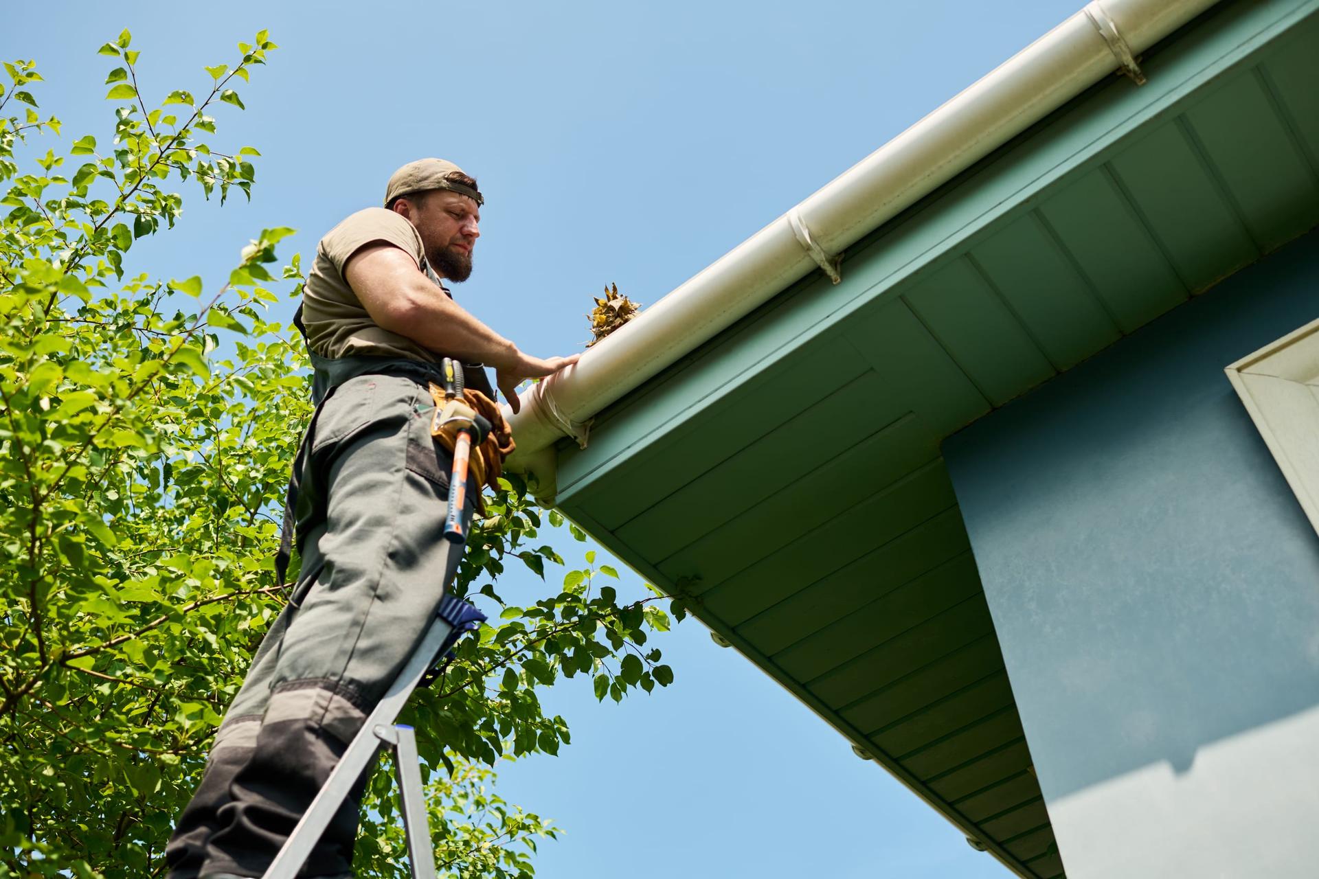 A man standing on a ladder pulling a clump of debris out of the eavestrough along the roofline of a house.