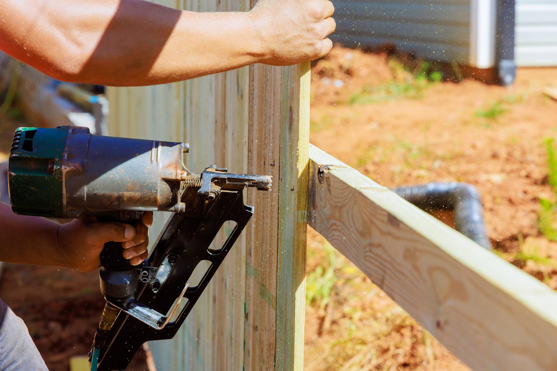 A worker's hands holding a pneumatic nail gun to fasten a pressure-treated rail to the post of a half-built wooden fence, with bare soil and house siding behind