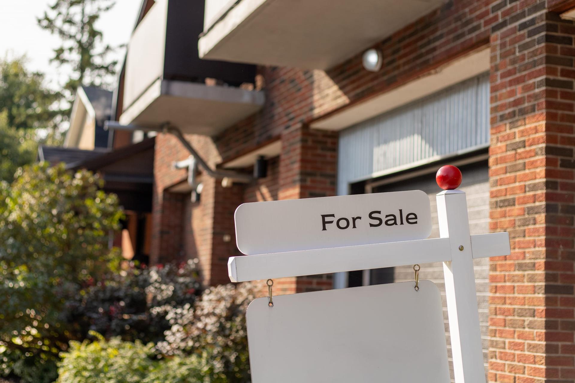A white For Sale sign on a post in front of a brick house with a garage door and balcony behind it.