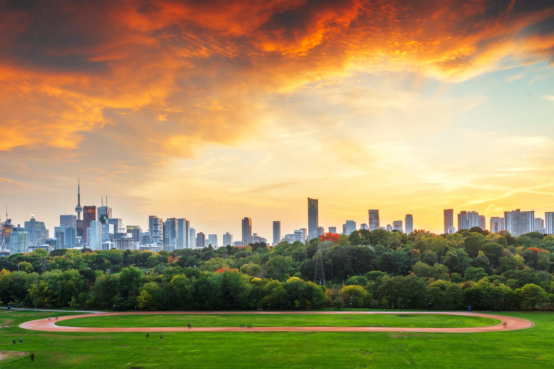 Toronto's downtown skyline and the CN Tower at sunset, seen across the grass and running track of a city park.