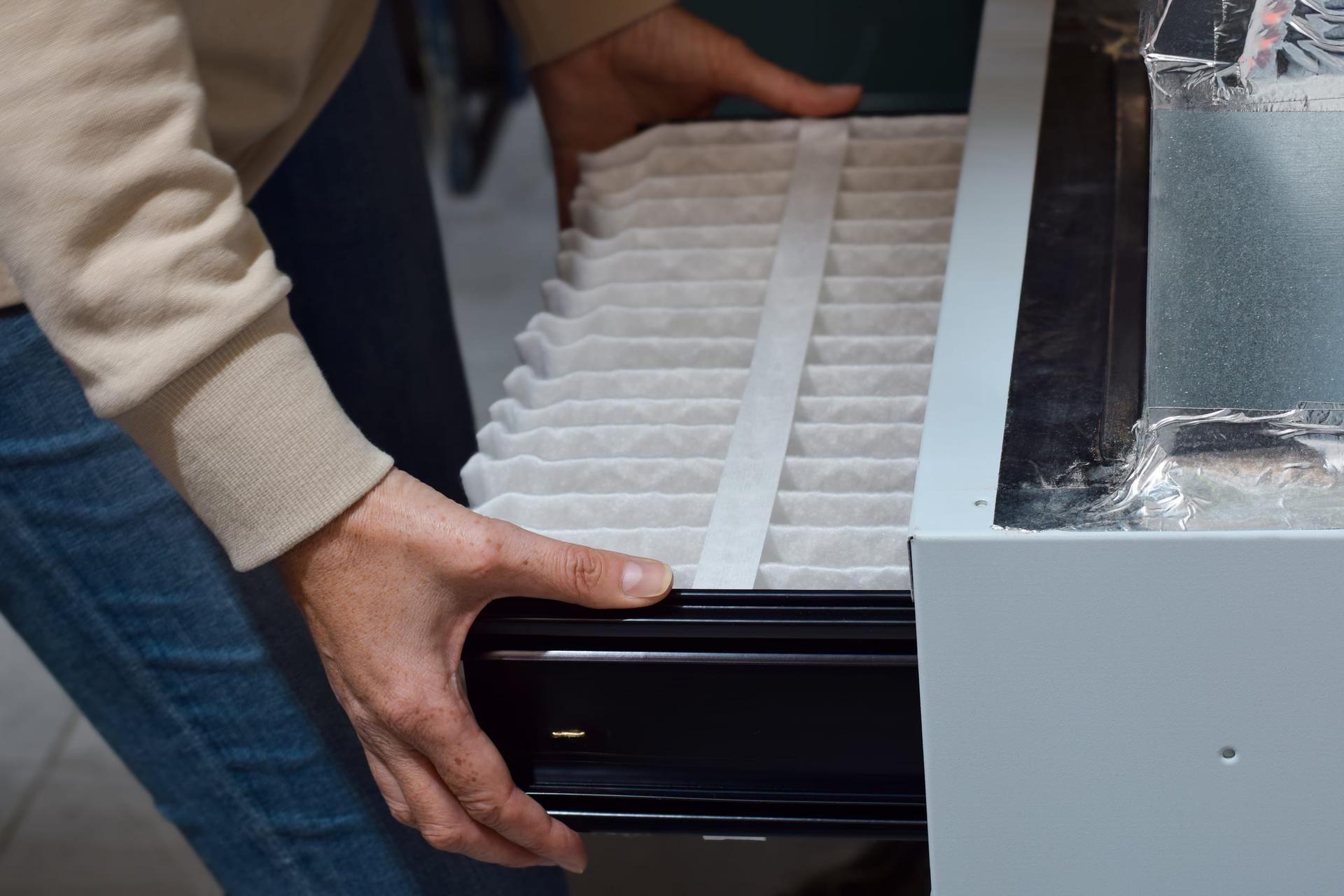 A person in a cream sweater and jeans sliding a white pleated filter into the filter slot of a white sheet-metal furnace cabinet