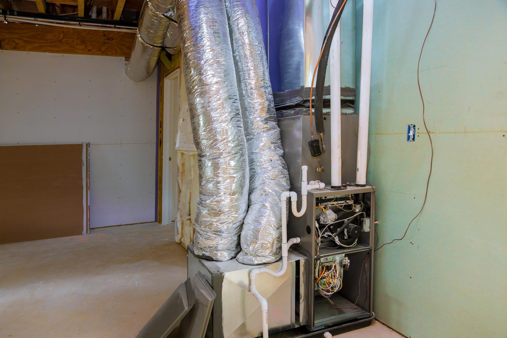 An upright gas furnace in an unfinished basement with its front panels removed, showing wiring and components inside the cabinet, white plastic vent and drain pipes beside it and silver insulated flex ducts rising from the plenum