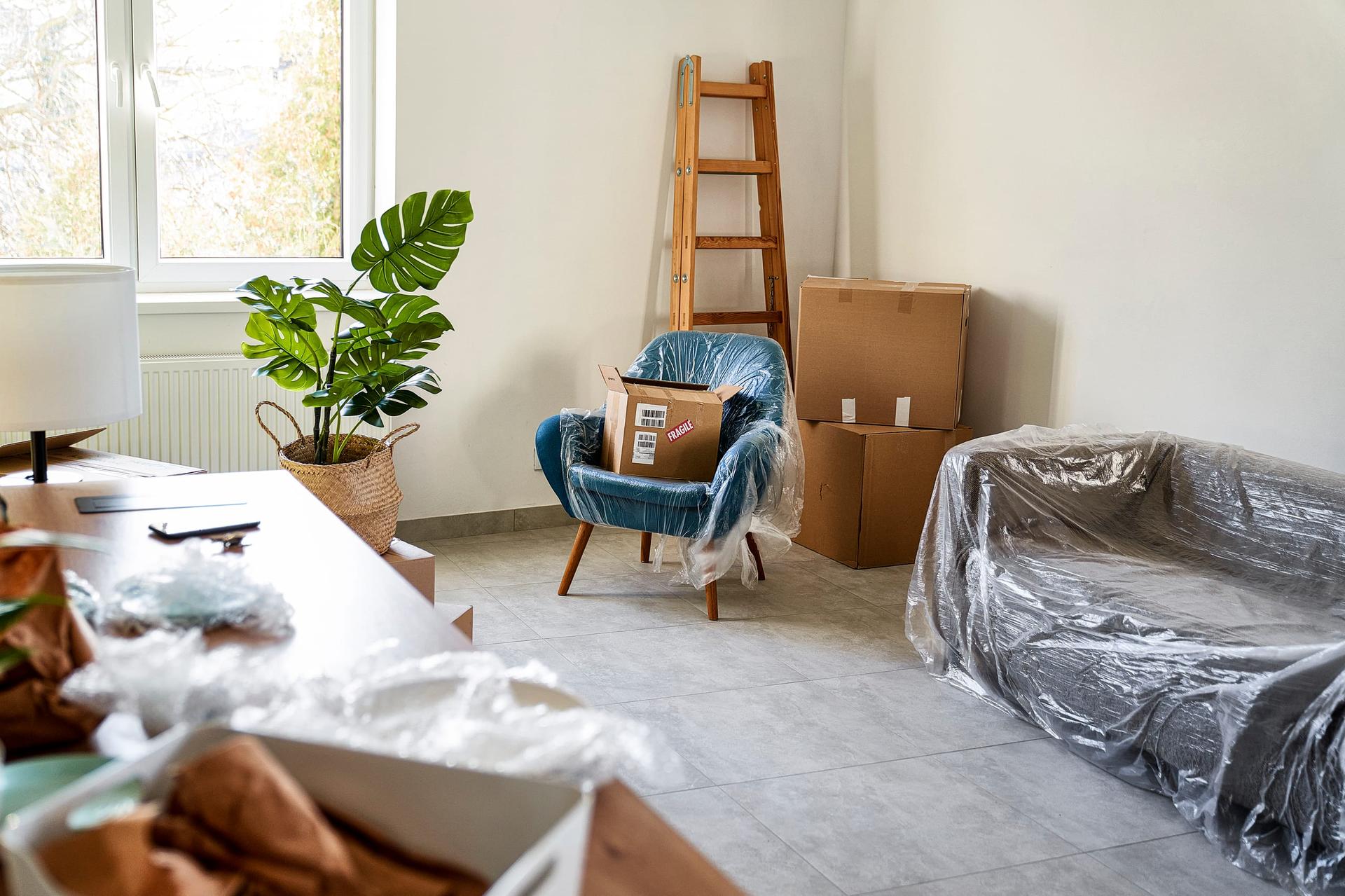 A half-packed living room with an armchair and sofa wrapped in plastic sheeting, stacked cardboard boxes and a wooden ladder against the wall.