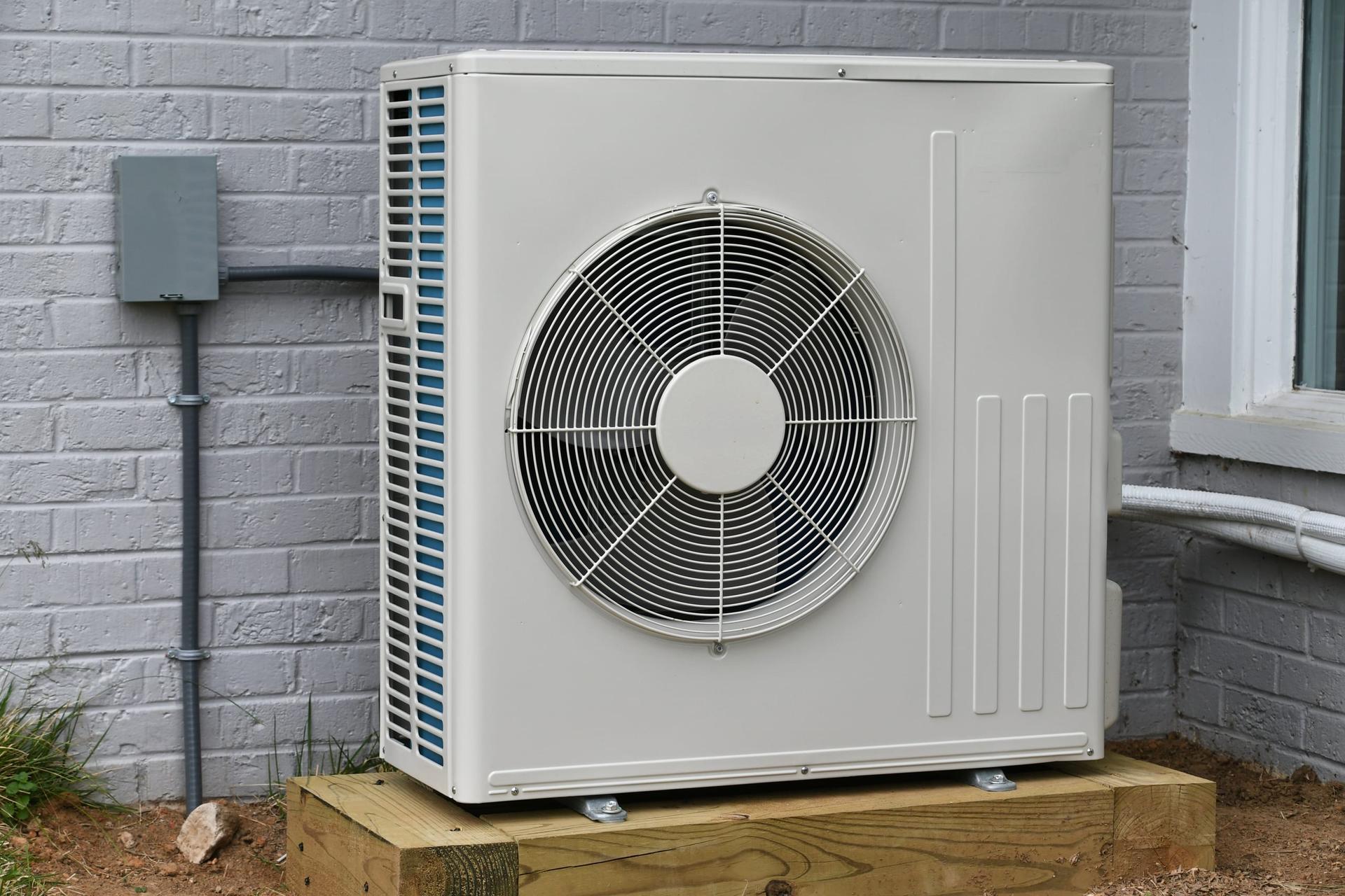 A white outdoor heat pump unit with a round fan grille on a timber pad against a grey-painted brick house wall, with an electrical disconnect box, conduit and a covered refrigerant line beside it