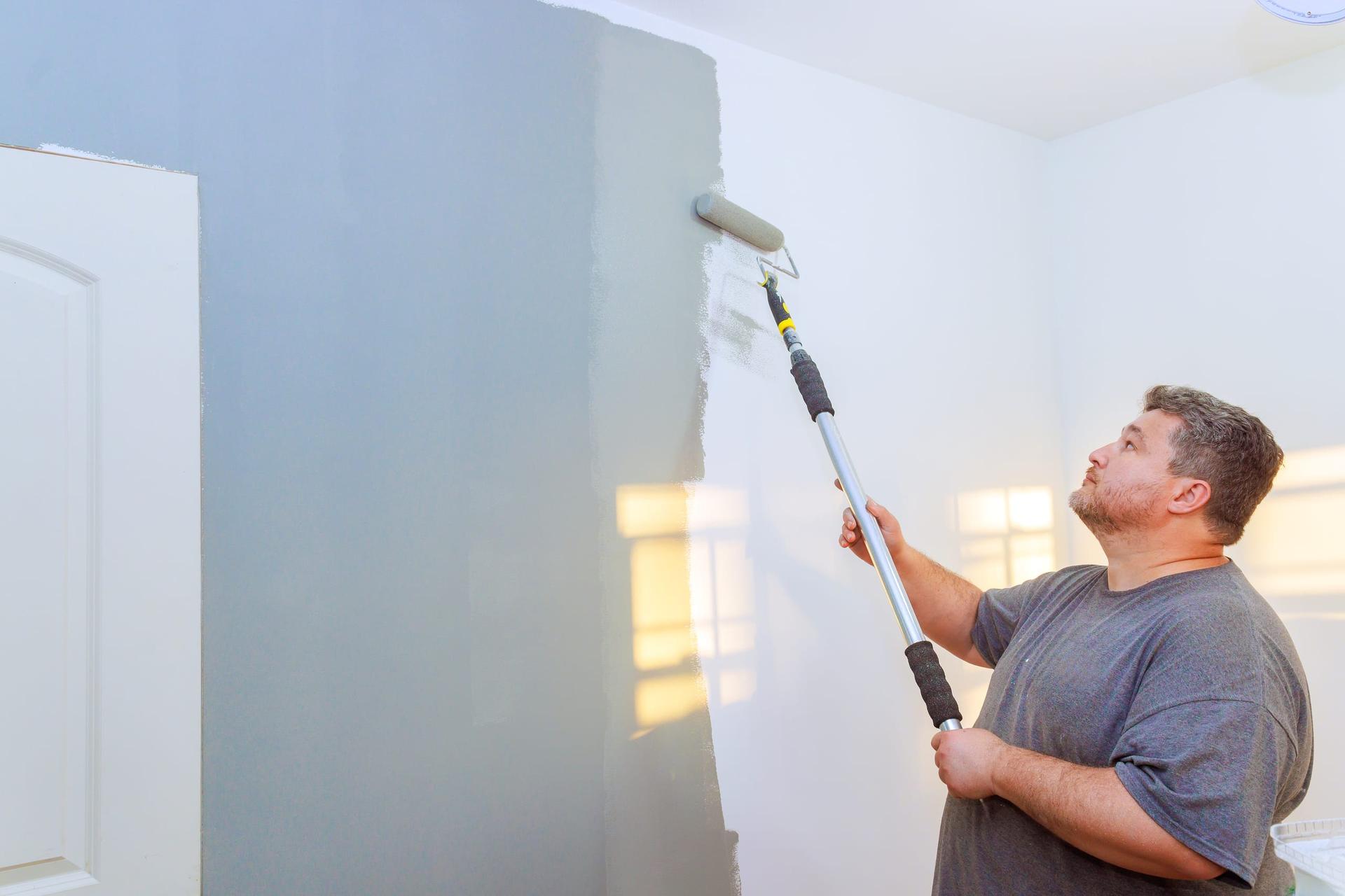 A man in a finished room using a roller on an extension pole to paint an interior wall, working along a wet edge beside a white panelled door with a sunlit window behind him
