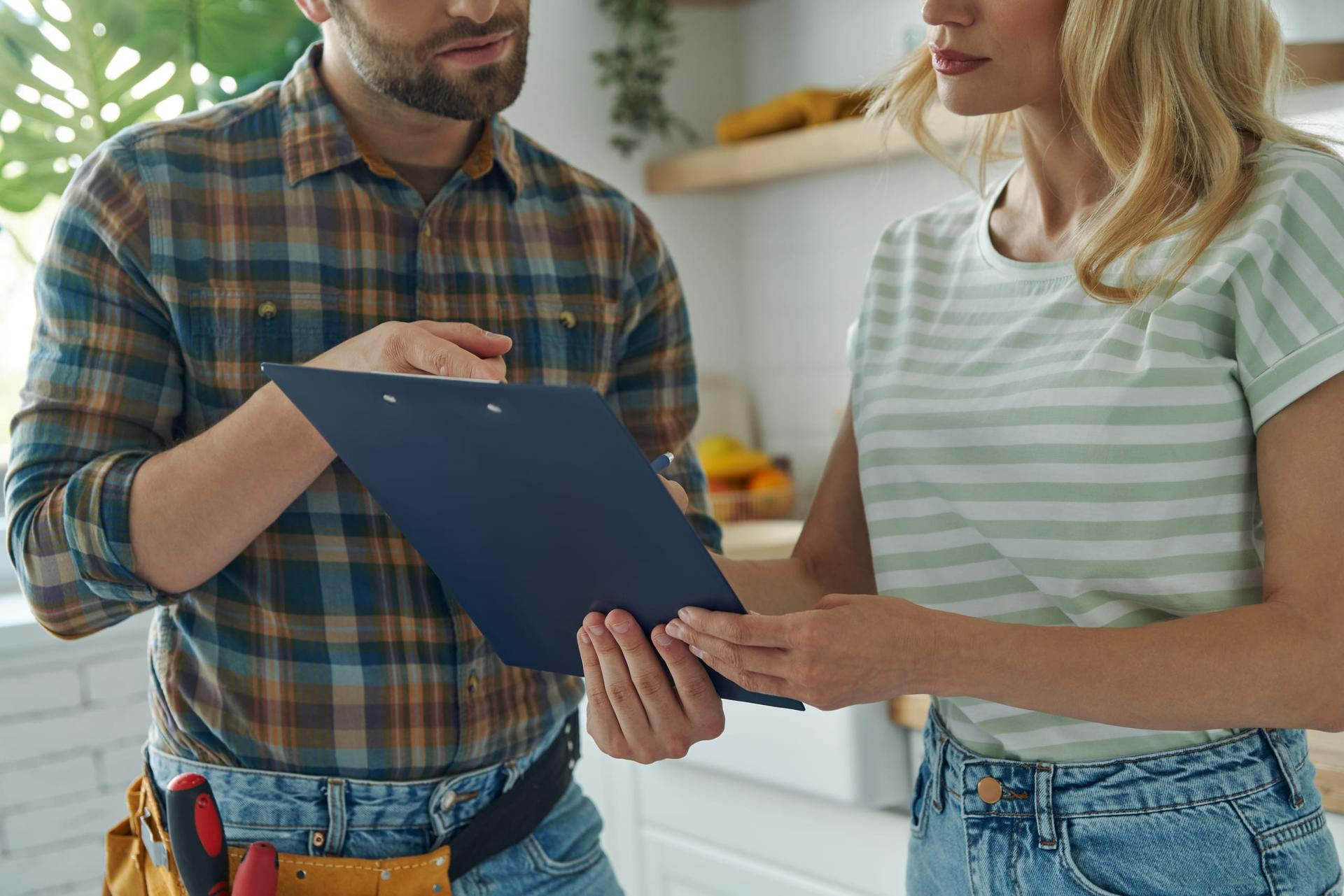 A tradesperson holds out a clipboard while a customer signs the paperwork clipped to it.