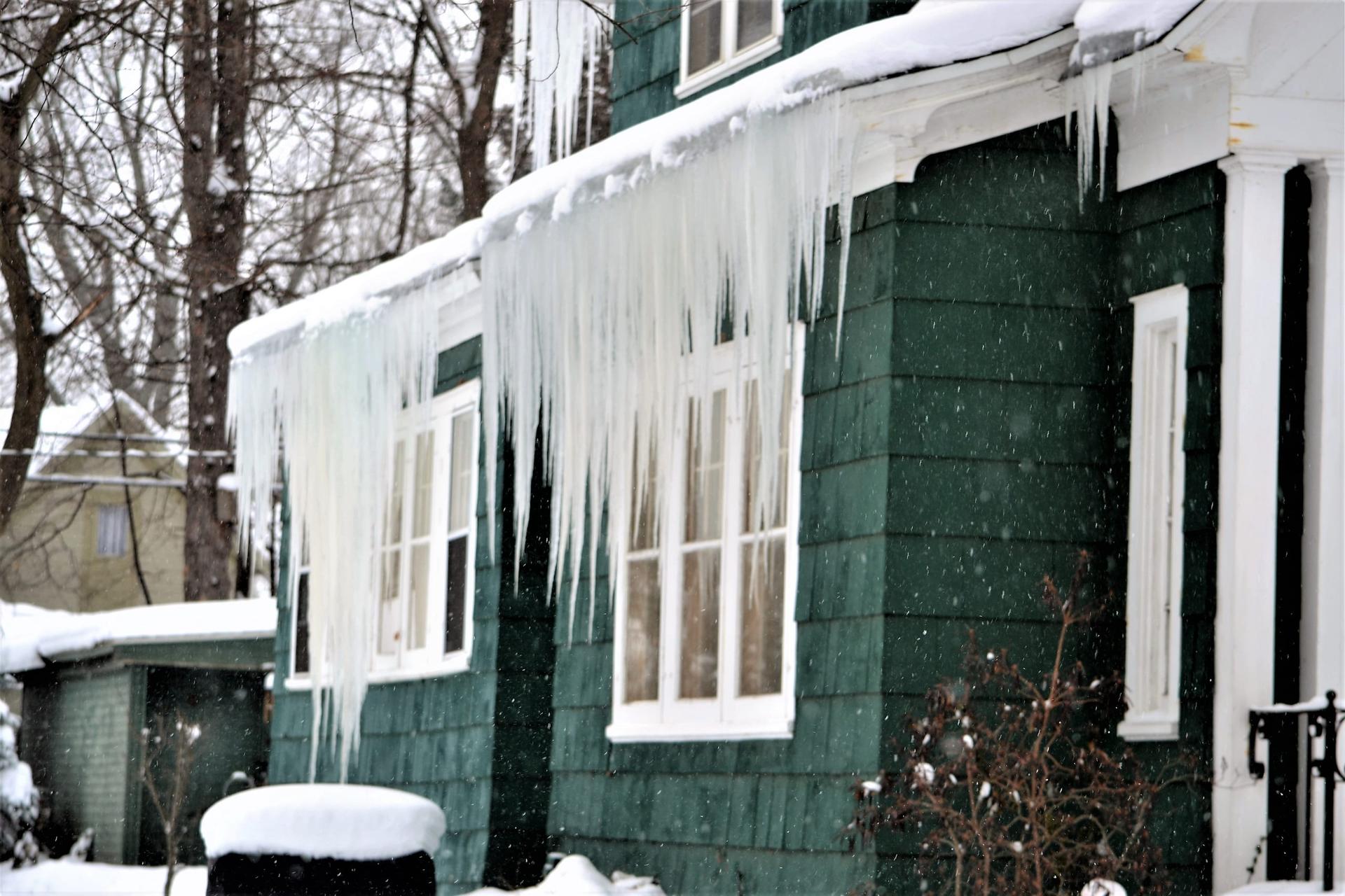 Long icicles hang in a thick curtain from the snow-covered eaves of a house with dark green shingle siding and white-framed windows, with bare trees behind and snow falling