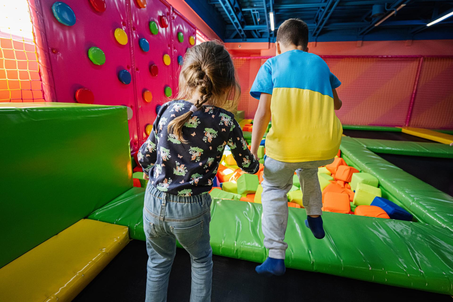 Two children seen from behind, a girl with a braid and a boy in blue socks, stepping onto a padded green ledge beside a pit of foam cubes and a pink climbing wall in an indoor play centre