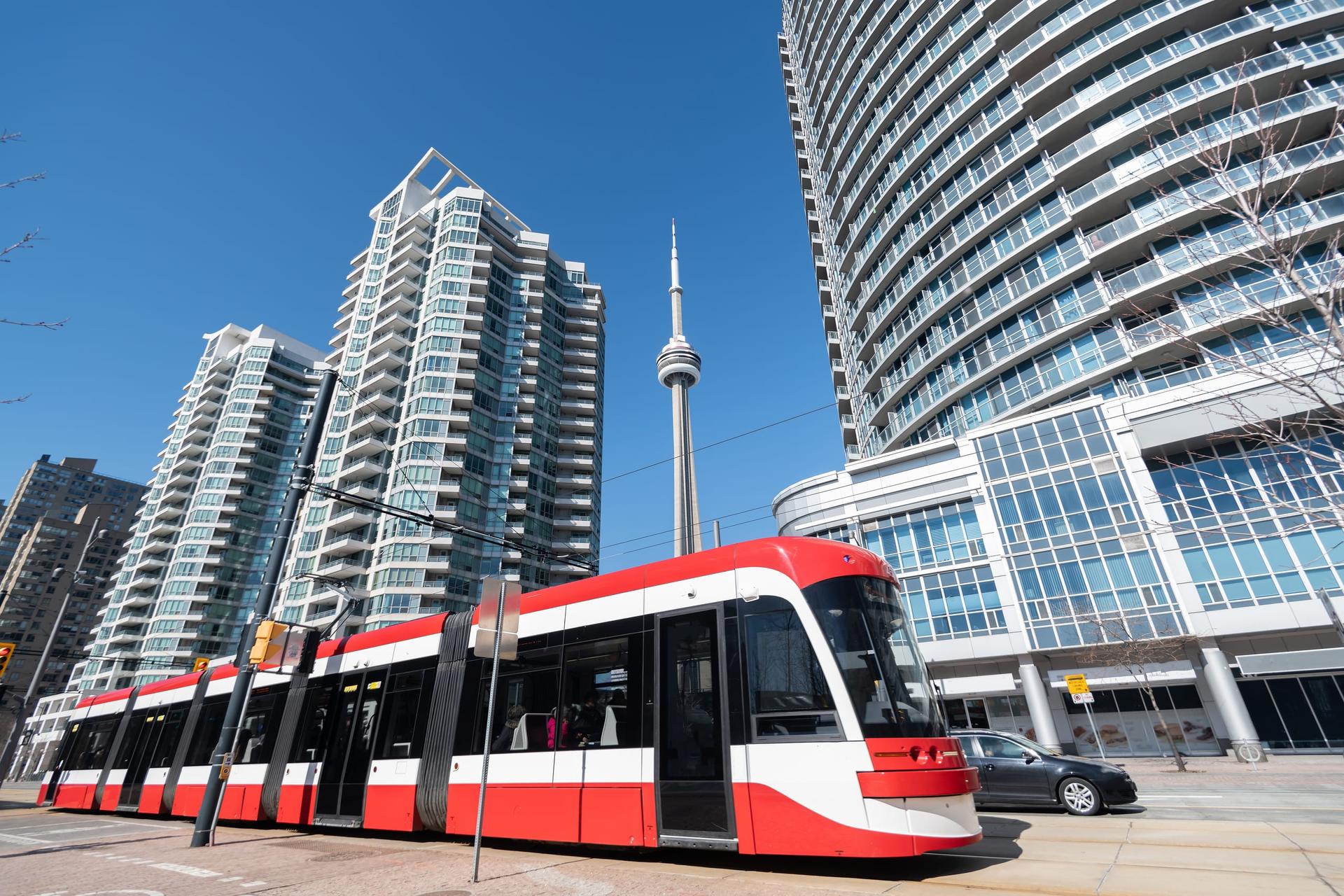 A red and white TTC streetcar on a sunny Toronto street with condo towers and the CN Tower behind it