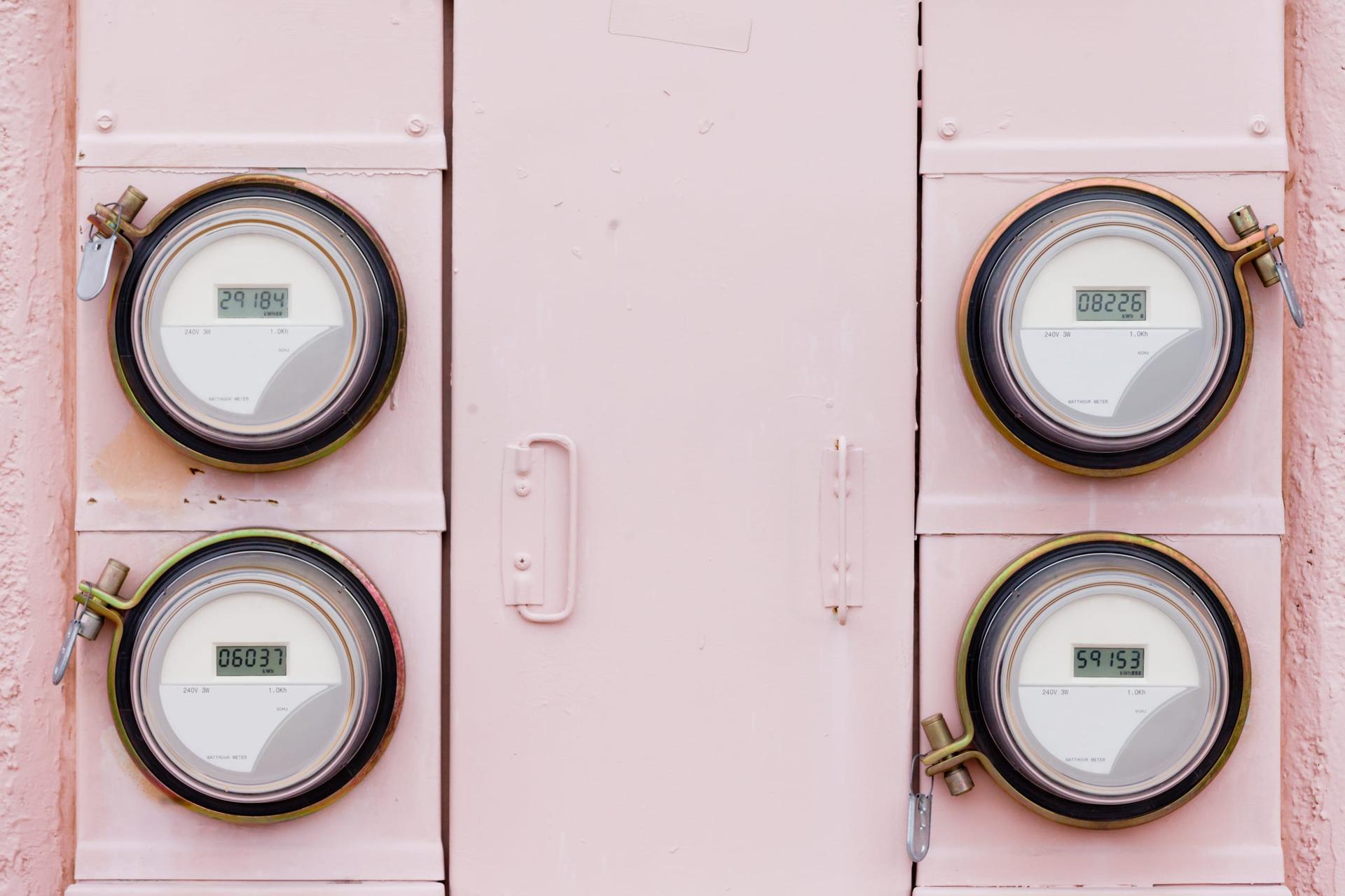 Four round digital electricity meters with kWh readouts, mounted in pairs on a pink metal meter cabinet on either side of a central access door