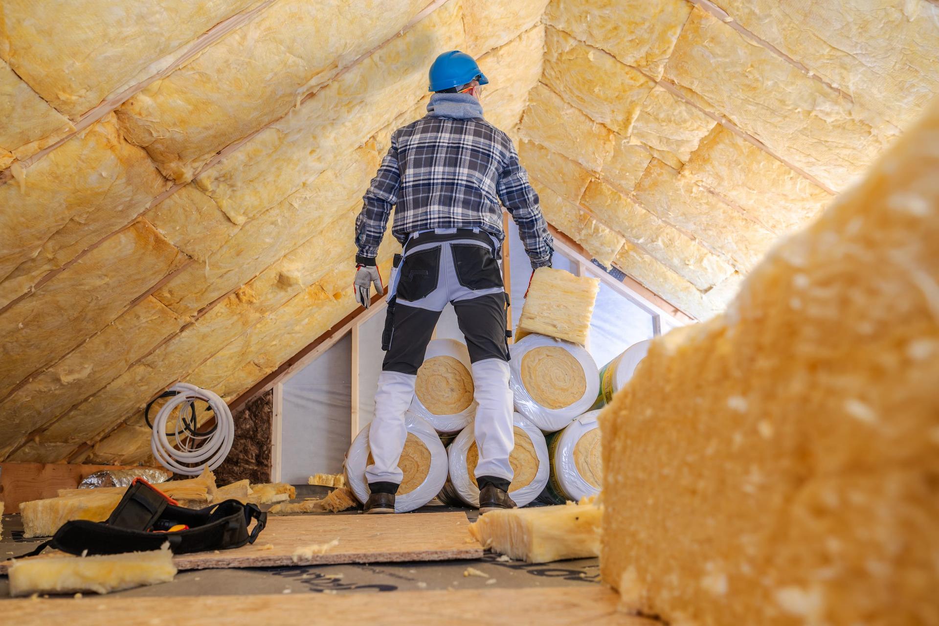 A worker in a blue hard hat, seen from behind, standing in a house attic in front of stacked rolls of yellow fibreglass insulation