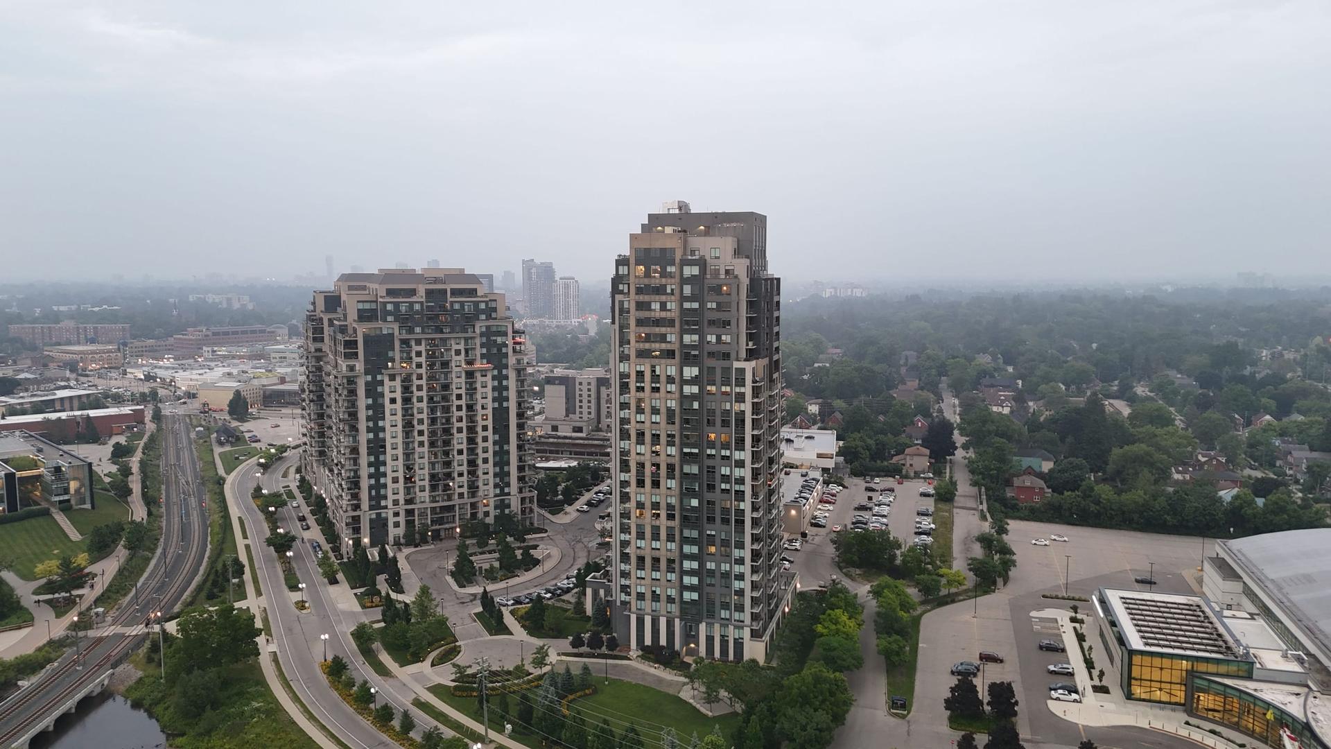 Aerial view of two high-rise apartment towers with balconies standing among low-rise houses and parking lots on an overcast day.
