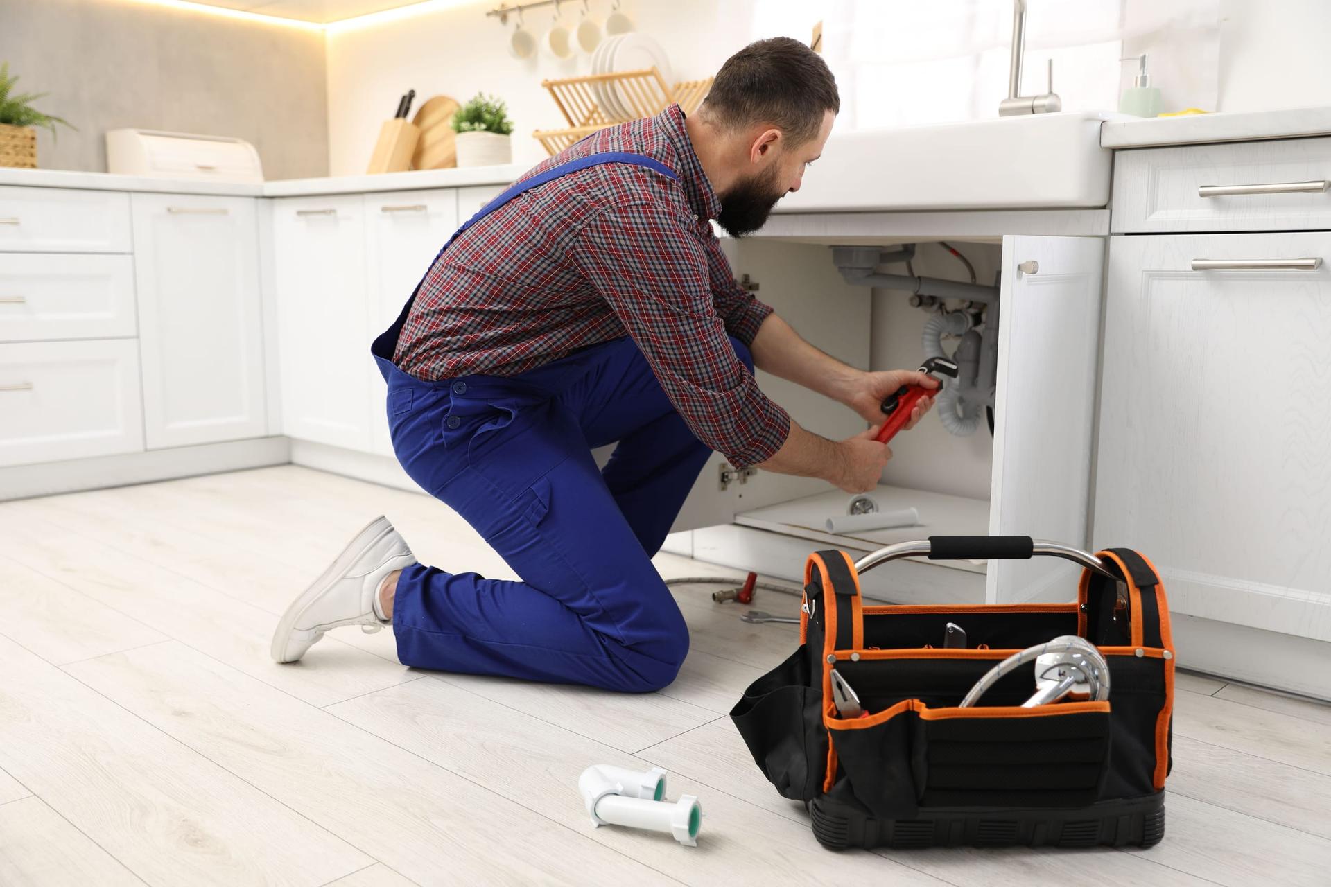 A plumber in blue overalls kneeling at an open under-sink cabinet, gripping the drain trap with a red-handled pipe wrench