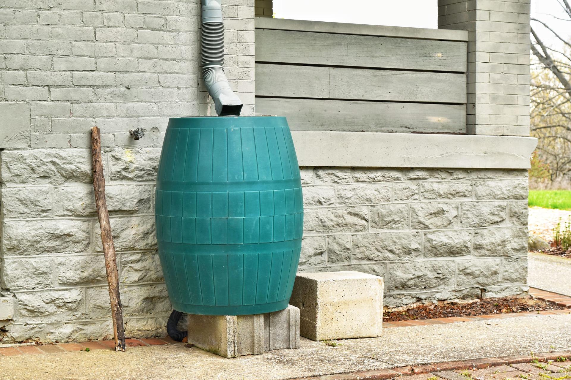 A teal plastic rain barrel raised on concrete blocks under a flexible downspout extension, against a grey-painted brick and stone house wall beside a porch