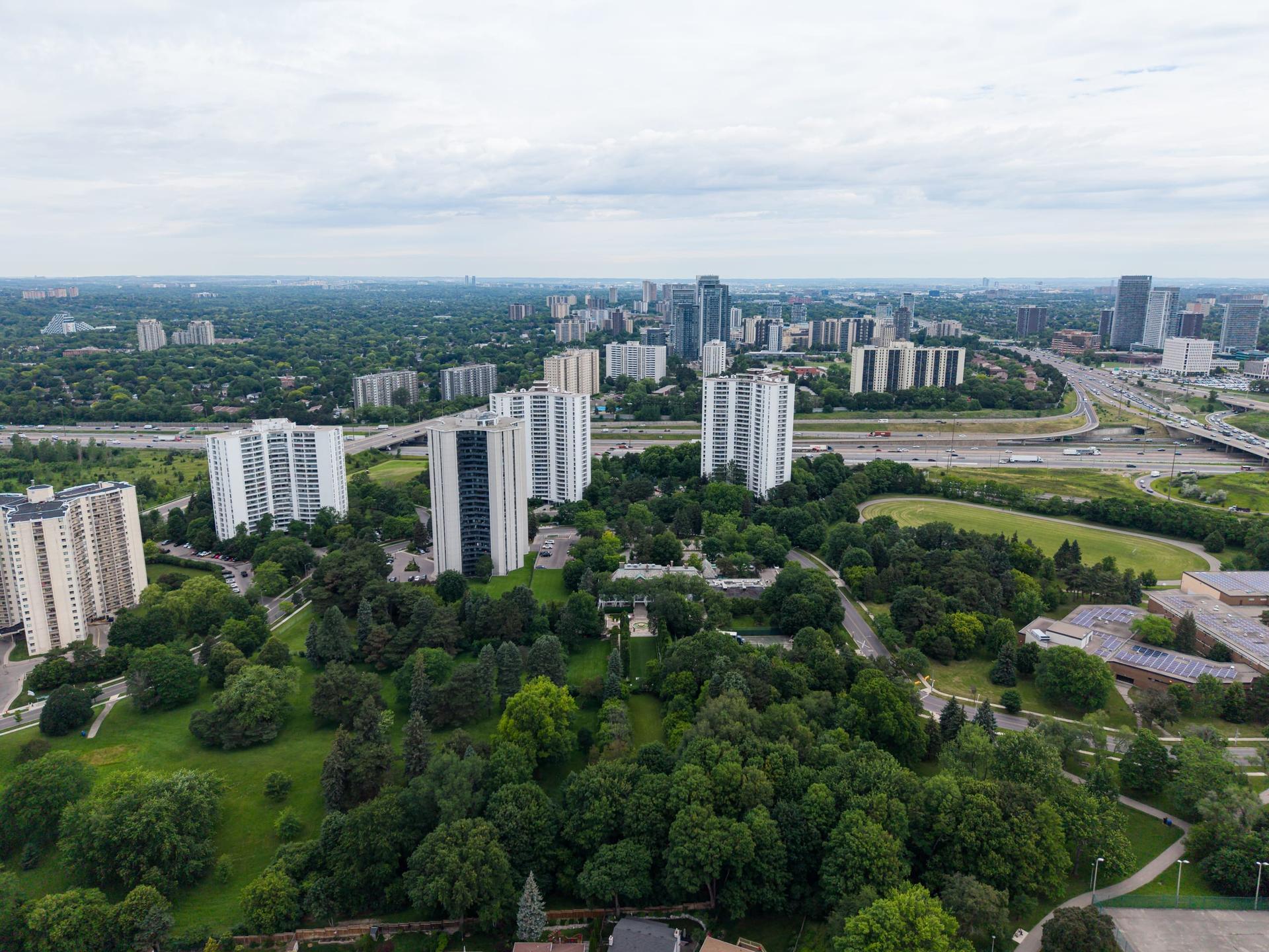 Aerial view of white and grey slab apartment towers among trees and parkland in North York, with Highway 401 and an interchange behind and more towers on the horizon under an overcast sky