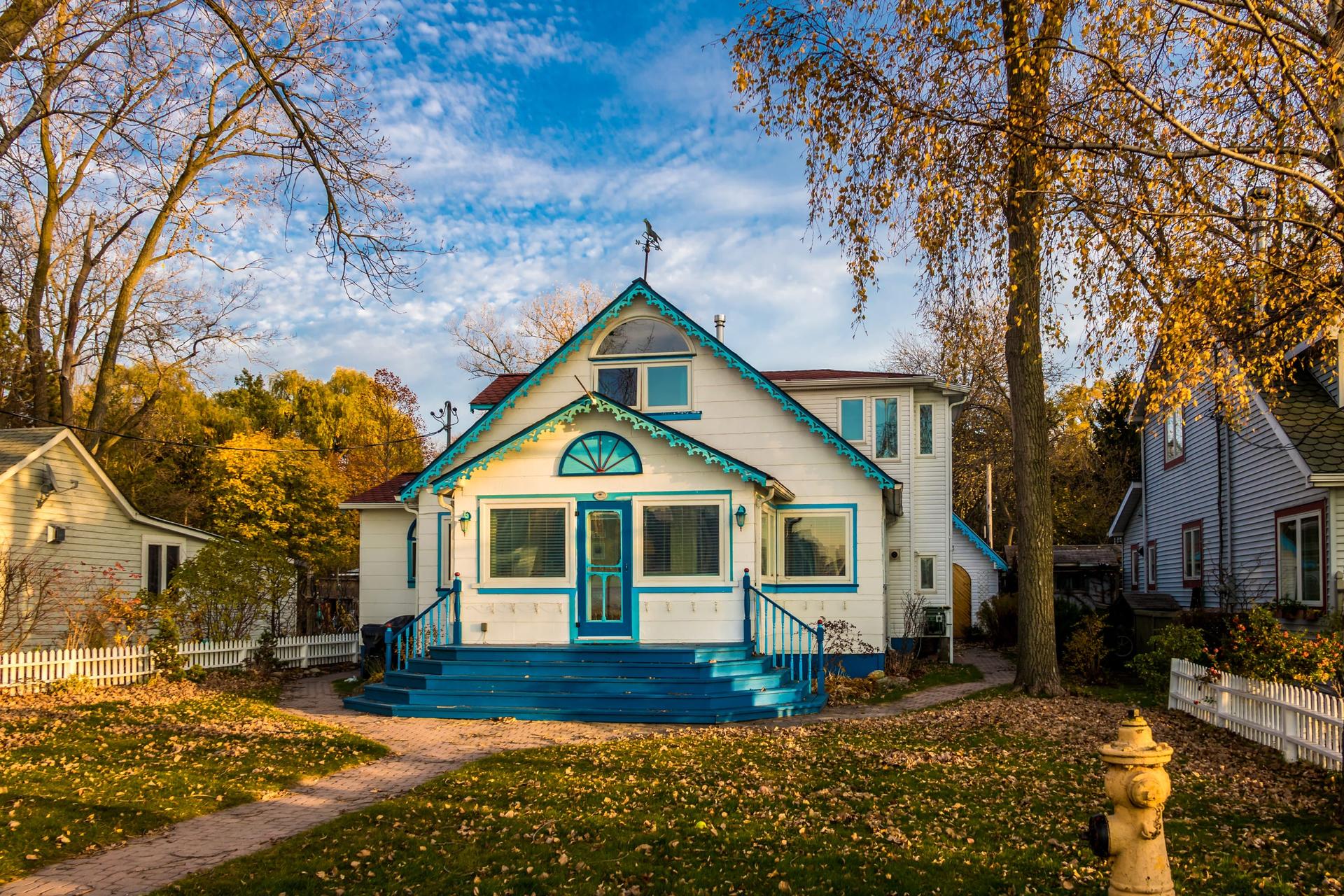 A white clapboard house with turquoise trim and blue porch steps behind a picket fence on a leafy Toronto street