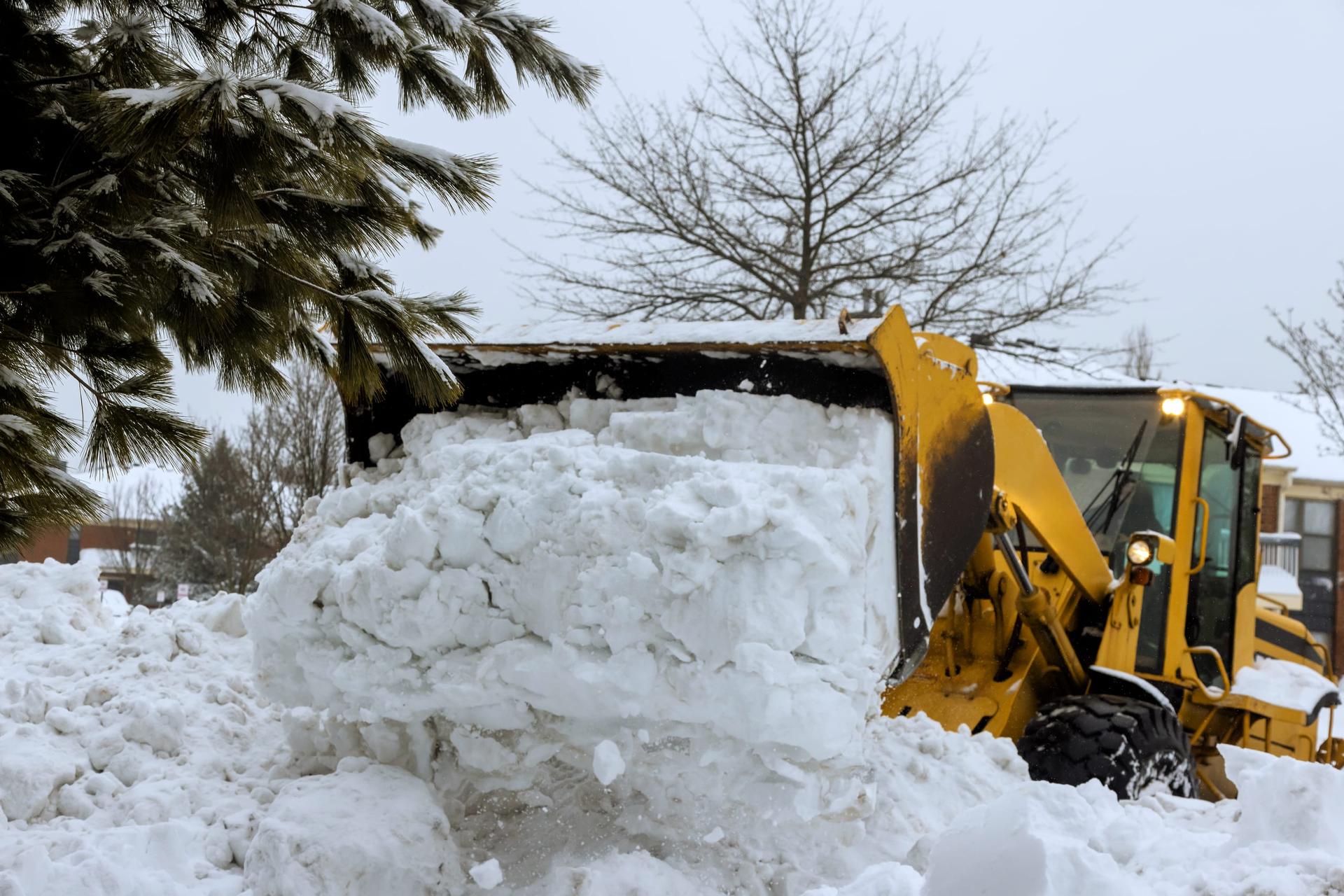 A yellow snow plow pushes a bank of deep snow along a residential street during heavy snowfall.