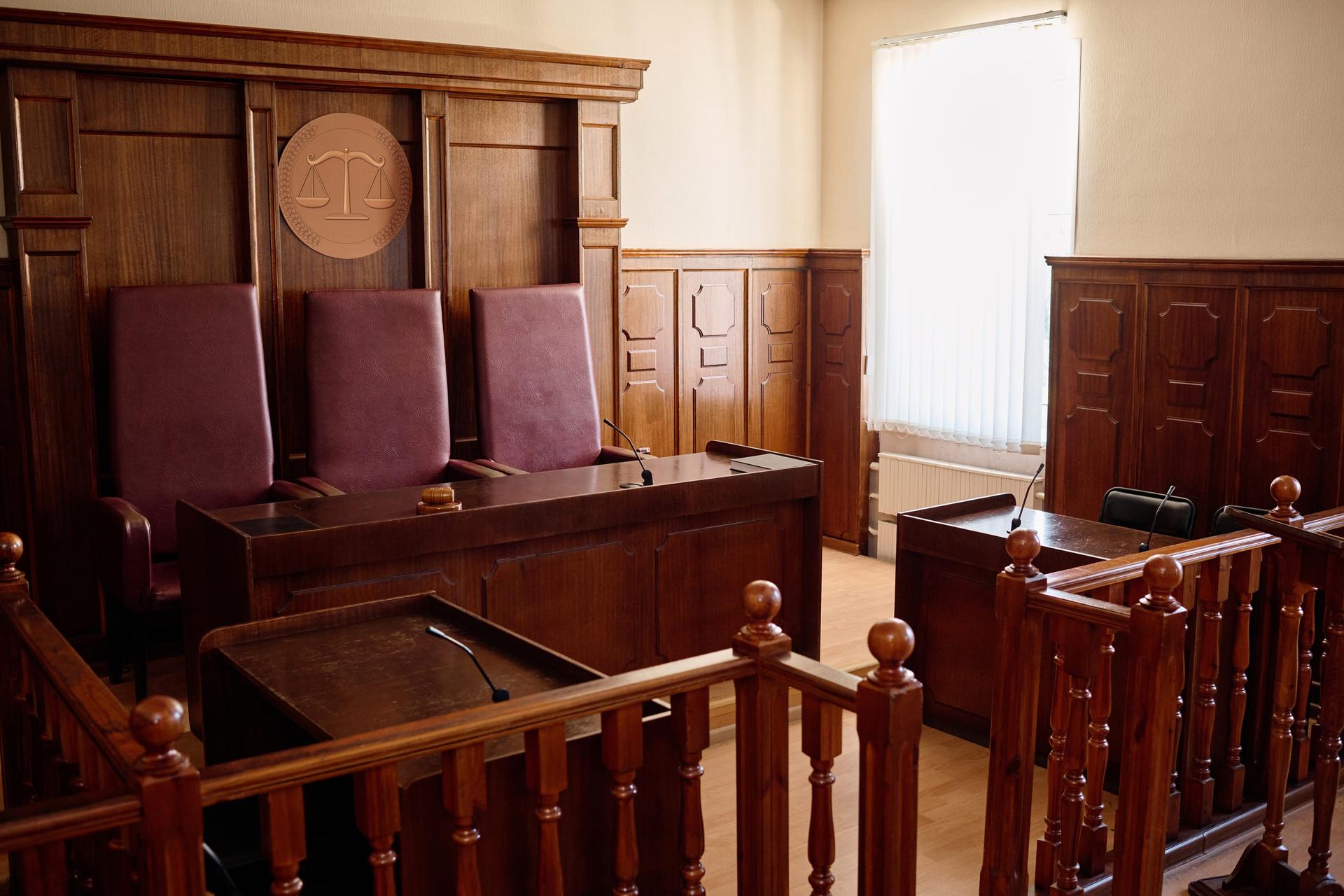 An empty wood-panelled hearing room with three high-backed chairs behind a raised bench and a wooden railing in the foreground.