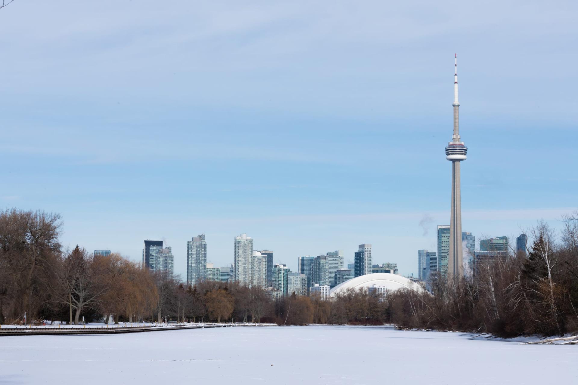 A snow-covered frozen lagoon on Toronto Island, with bare trees along the shore and the downtown skyline, the CN Tower and a white stadium dome behind it under a pale blue sky