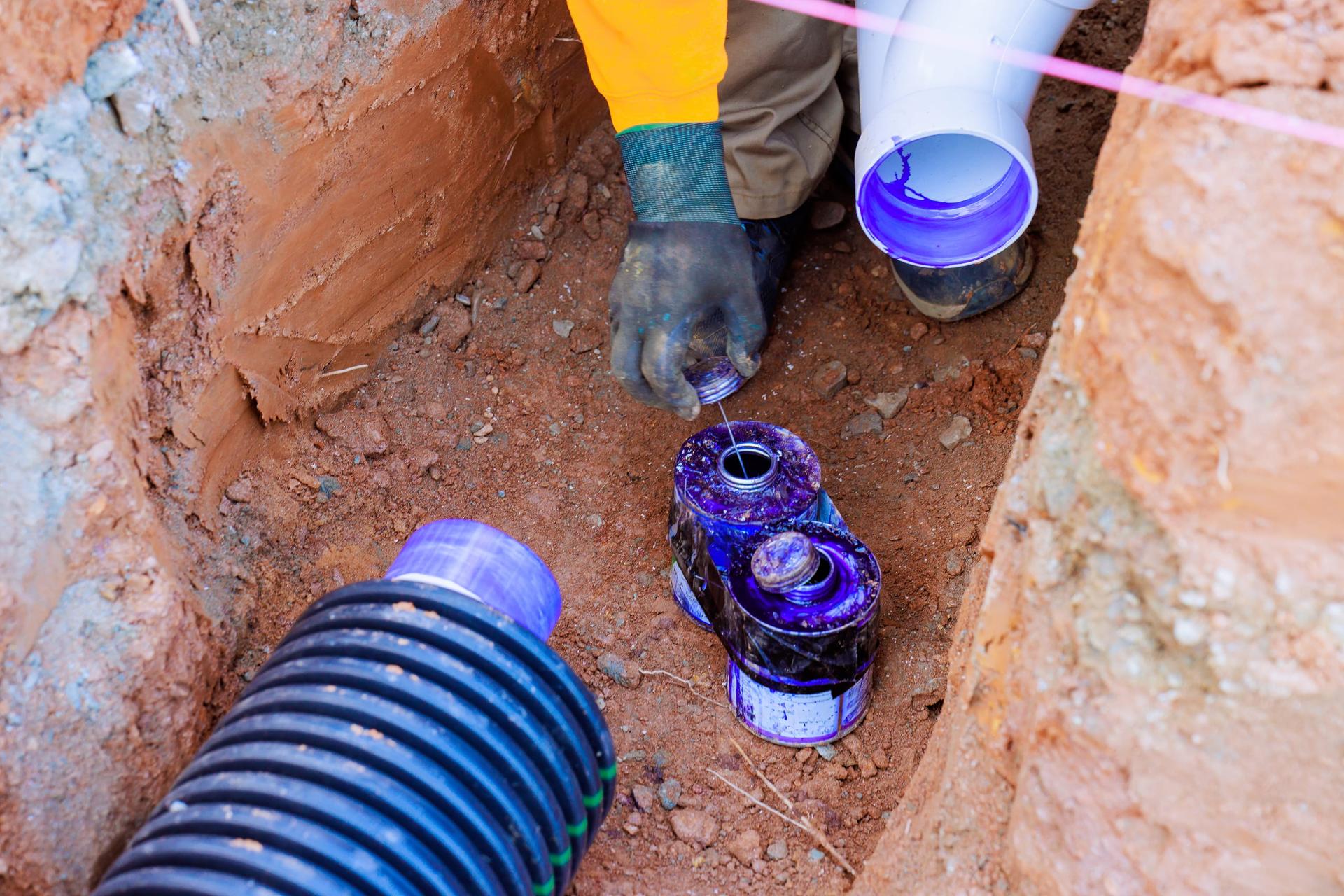A gloved hand in an open earth trench lifting the lid from a can of purple PVC primer, beside a freshly primed white drain fitting and a black corrugated pipe
