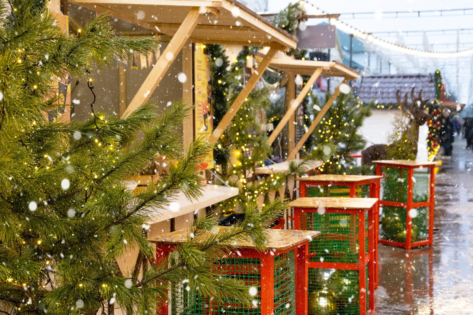 A row of wooden market stalls strung with fairy lights and evergreen boughs as snow falls at dusk.