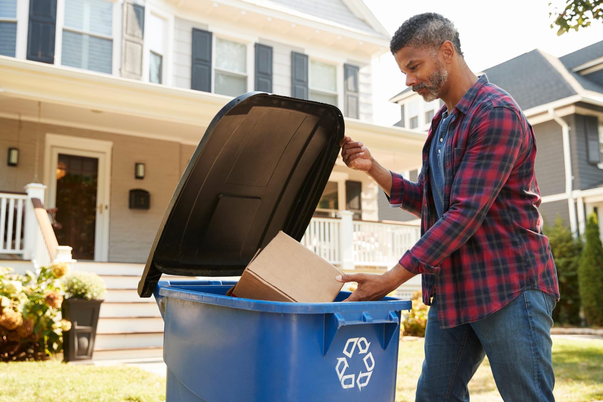 A man placing a cardboard box into a blue recycling bin in front of a house with a porch