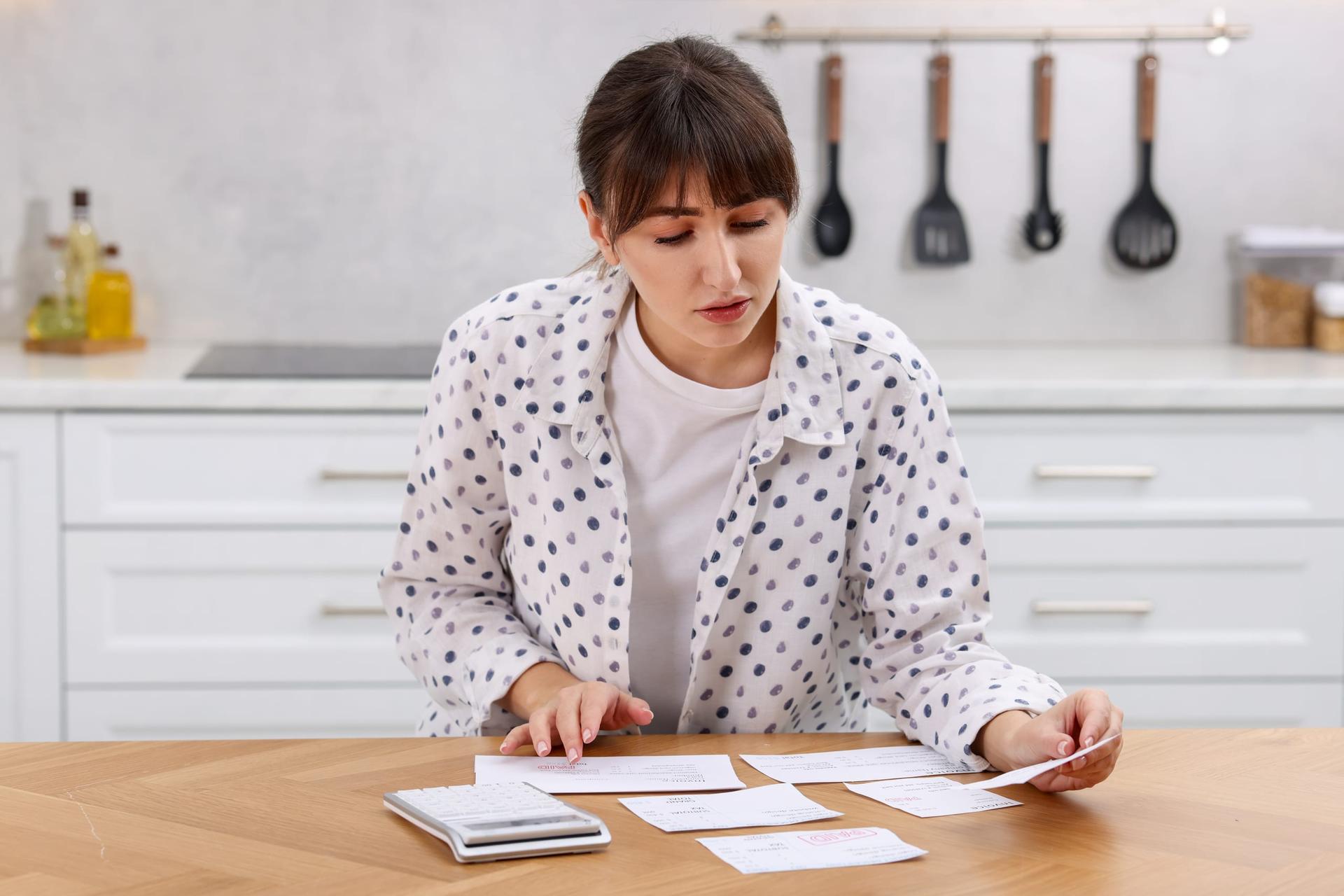 A person sits at a wooden table sorting through paper invoices with a calculator to hand.