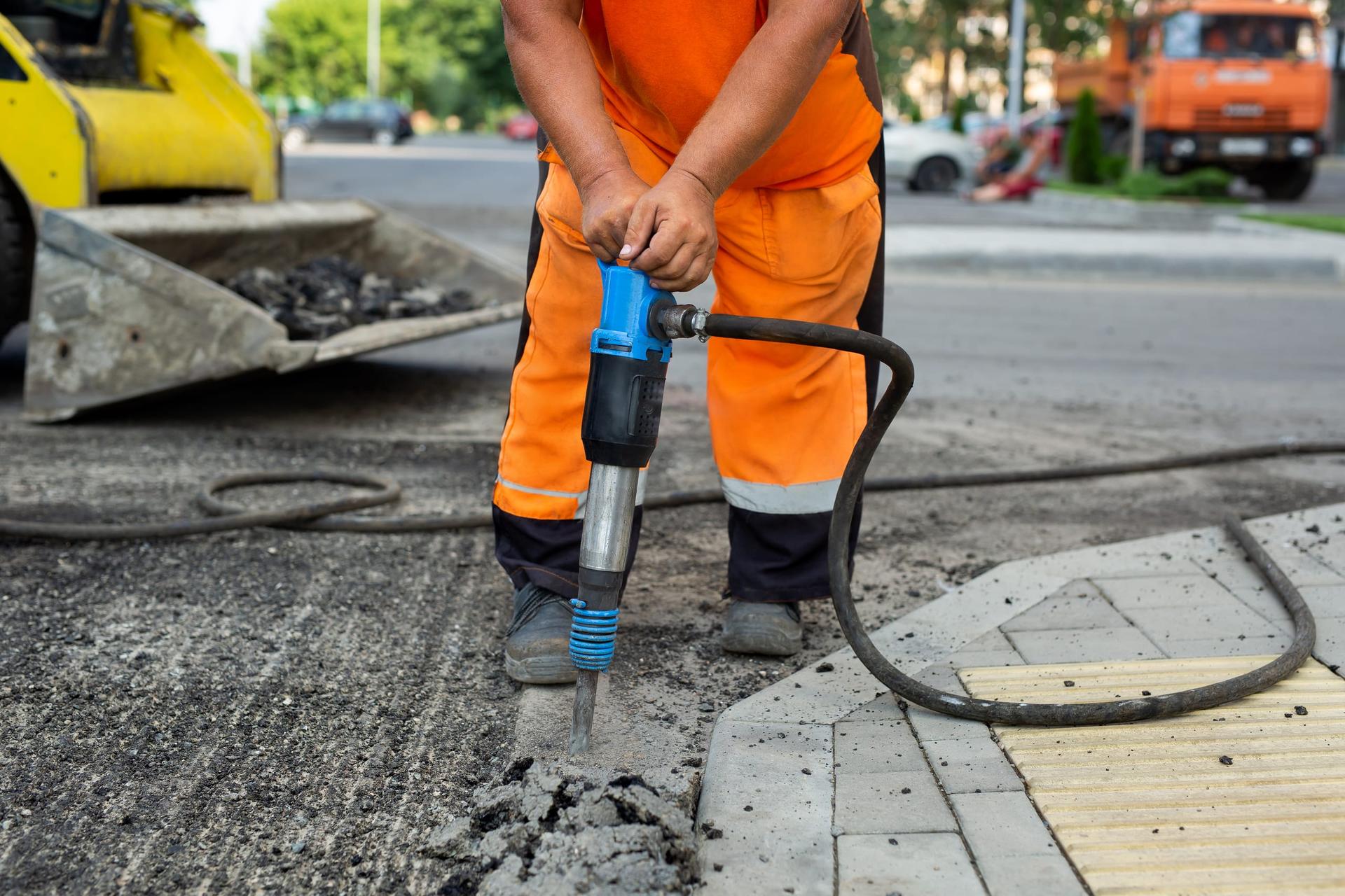 A road worker in an orange hi-vis suit breaking up street asphalt with a handheld jackhammer beside a paving-stone sidewalk.