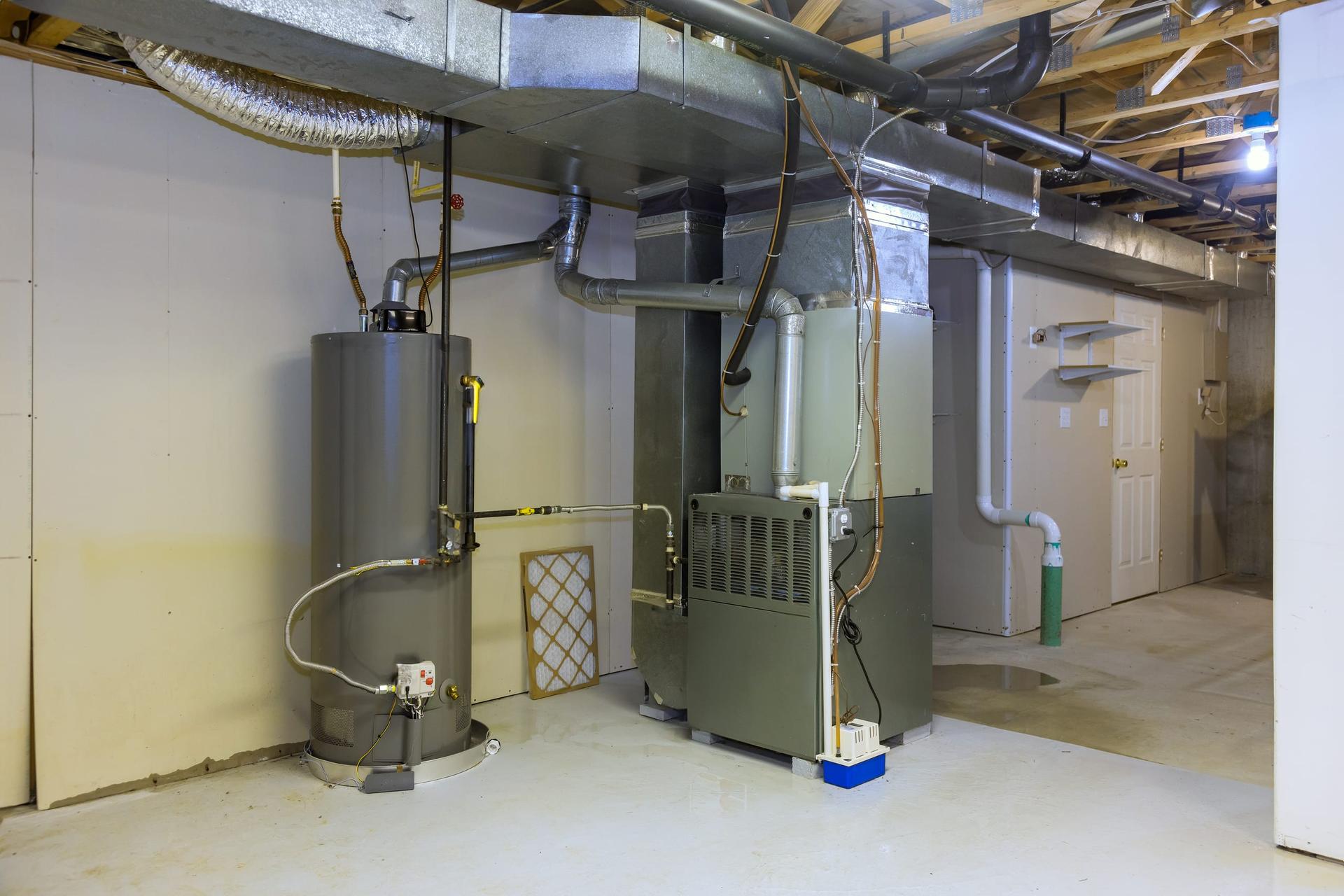 A tall gas water heater tank standing beside a furnace in an unfinished basement, with ductwork and vent pipes running overhead.