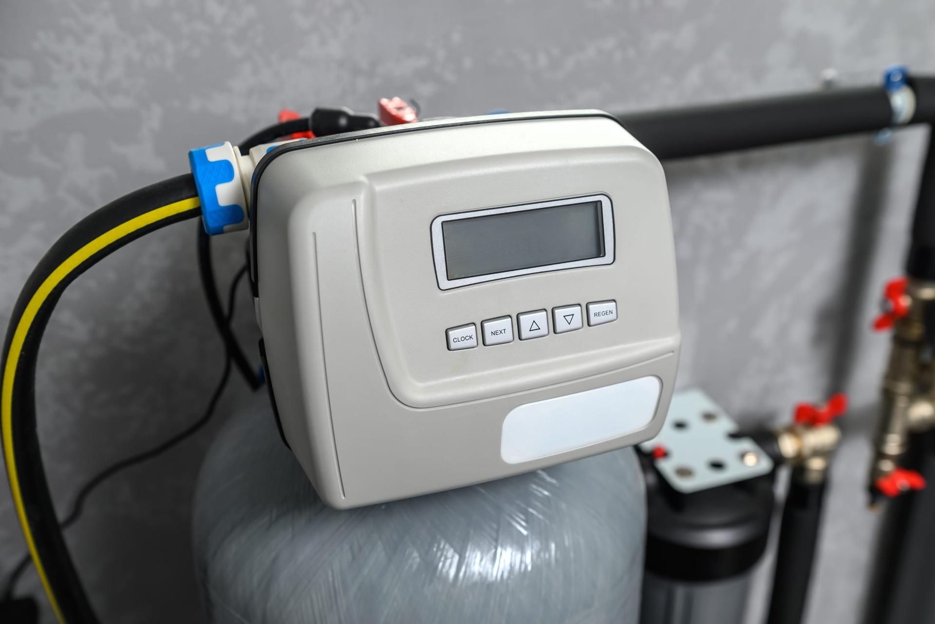 Close-up of a water softener's grey control valve, with a small LCD screen and CLOCK, NEXT, up, down and REGEN buttons, on top of a grey resin tank, with a black and yellow hose, a blue fitting, brass valves with red handles and a black filter housing beside it