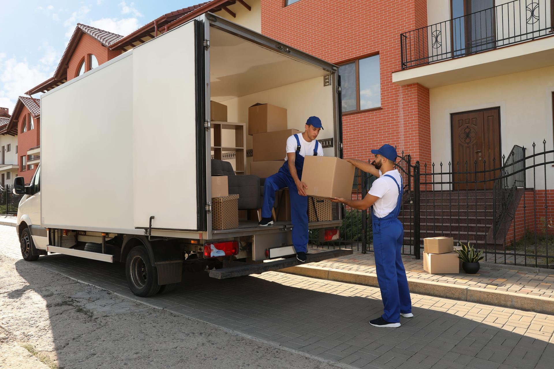Two movers in blue overalls pass a cardboard box out of the open back of a loaded white box truck parked outside a brick house.