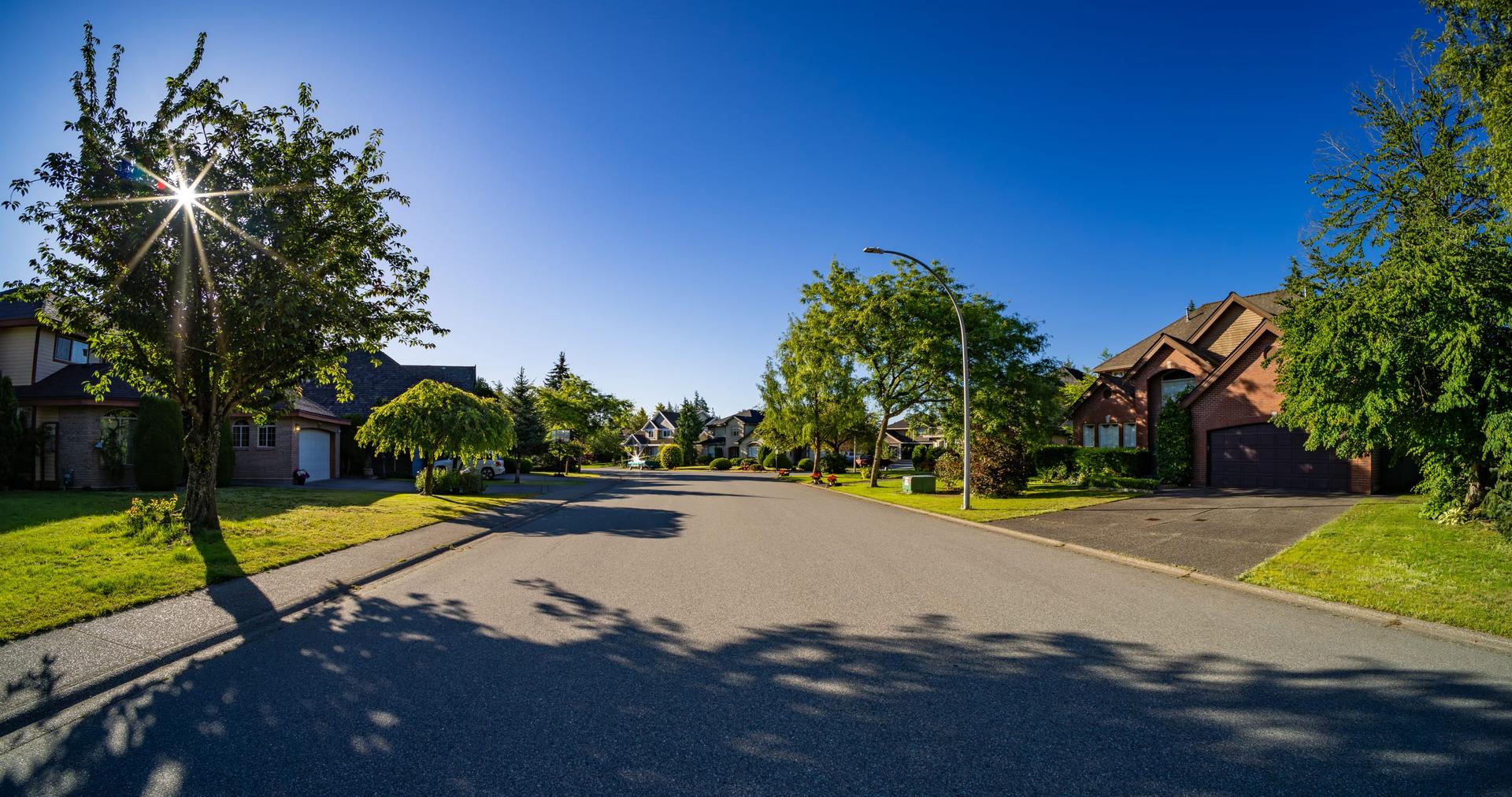 A sunlit suburban street of detached brick houses with attached garages and mown front lawns.