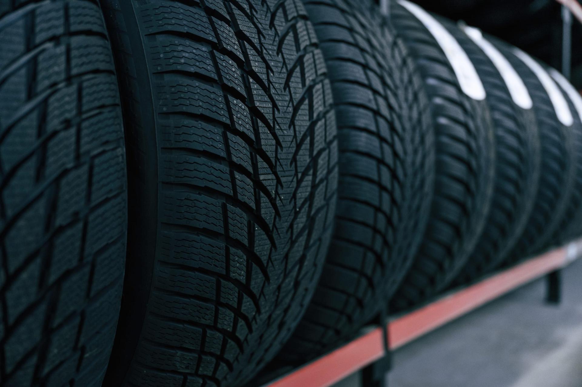 A row of black winter tires with deep V-shaped tread and fine slits stands upright on a red metal rack in a dim tire shop, the tires further back carrying white paper labels
