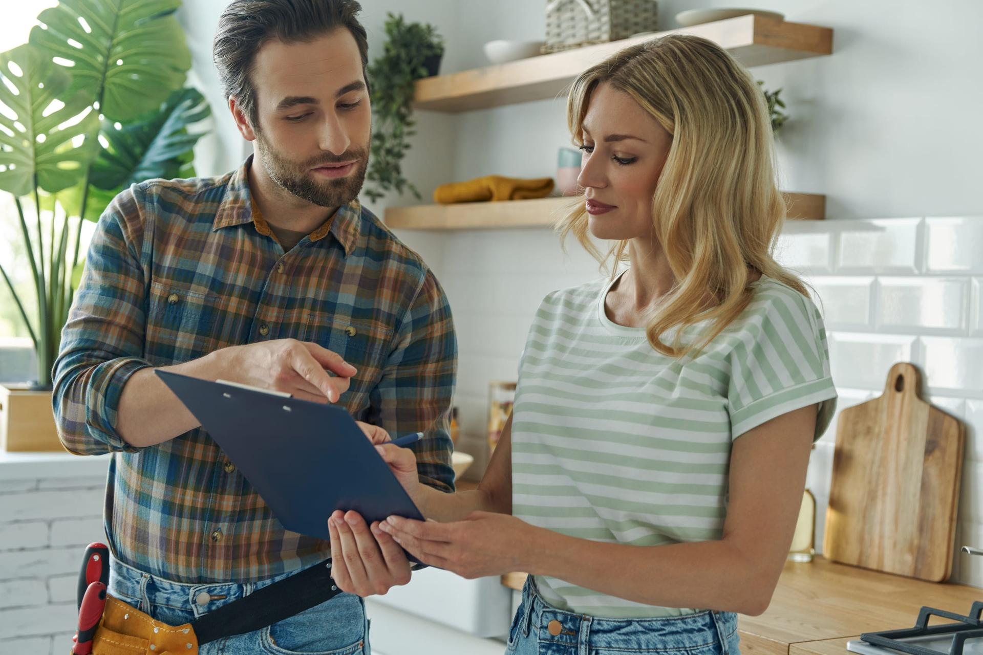 A tradesman in a plaid shirt and tool belt points to a page on a clipboard while a woman in a striped T-shirt reads it with a pen in hand, in a home kitchen